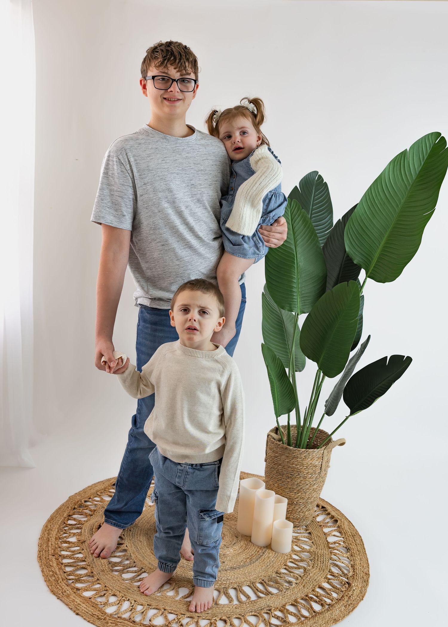 Teen boy holding toddler girl while young boy holds his hand, posing near tropical plant on woven rug.