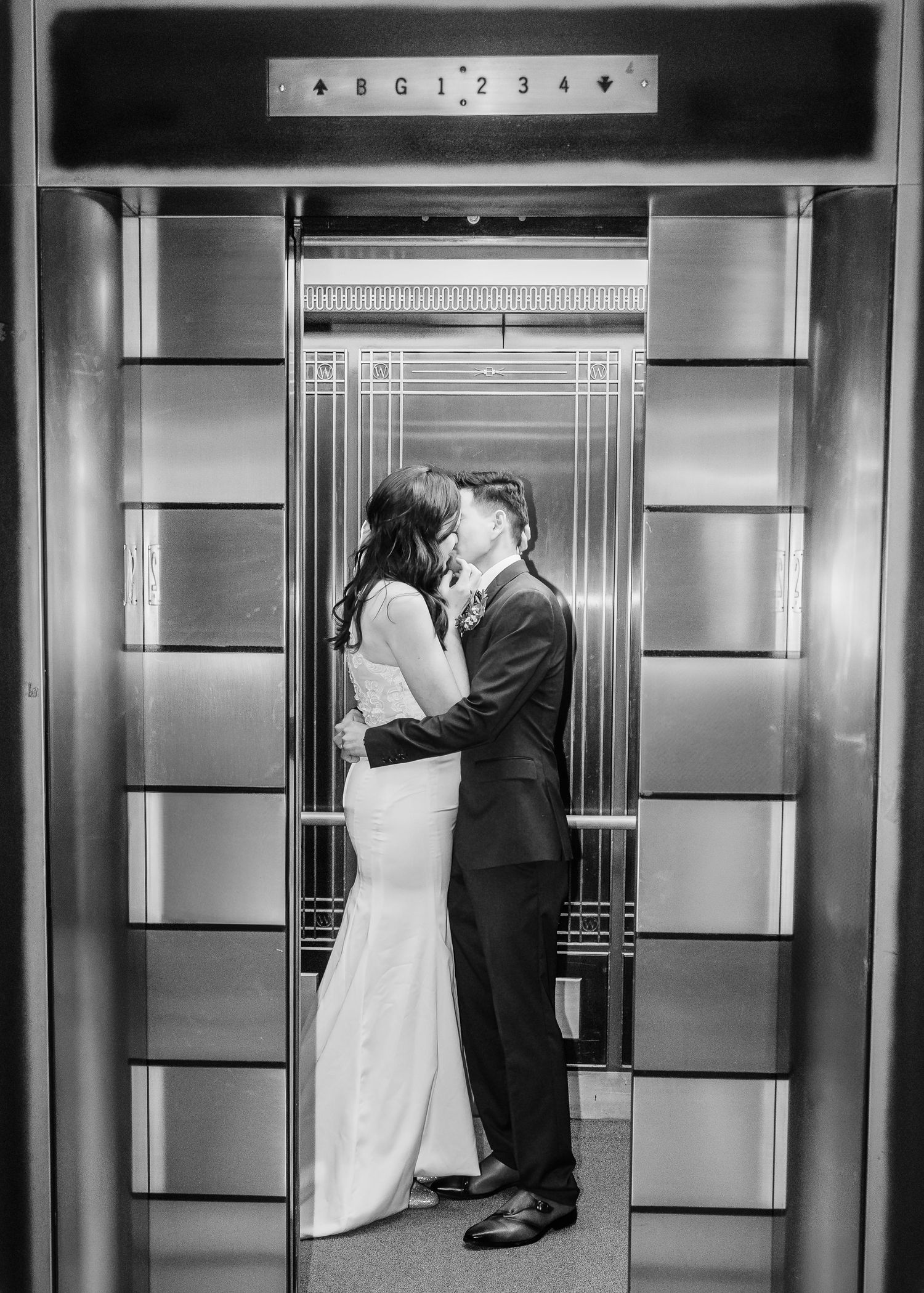 Black and white image of newlyweds kissing inside a vintage Art Deco elevator with decorative doors.