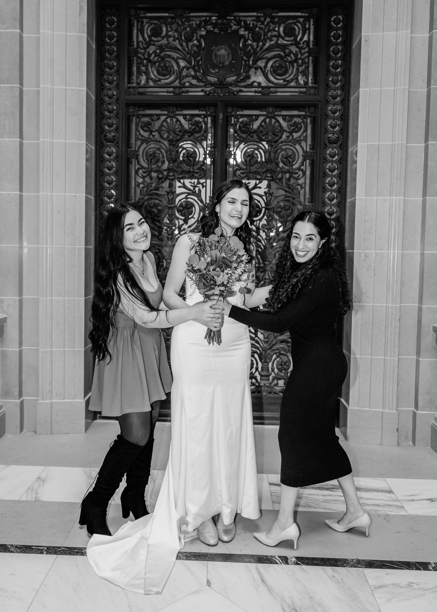 Bride in white gown holding bouquet flanked by two women before ornate doors, black and white.