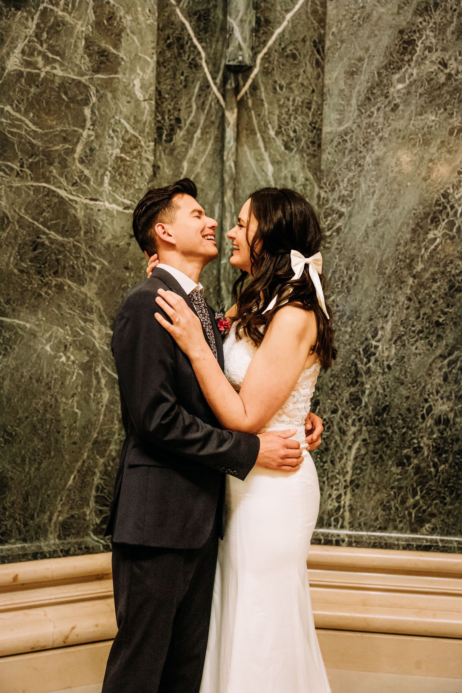 Smiling groom in dark suit embraces bride in white dress with bow, standing before marble wall.