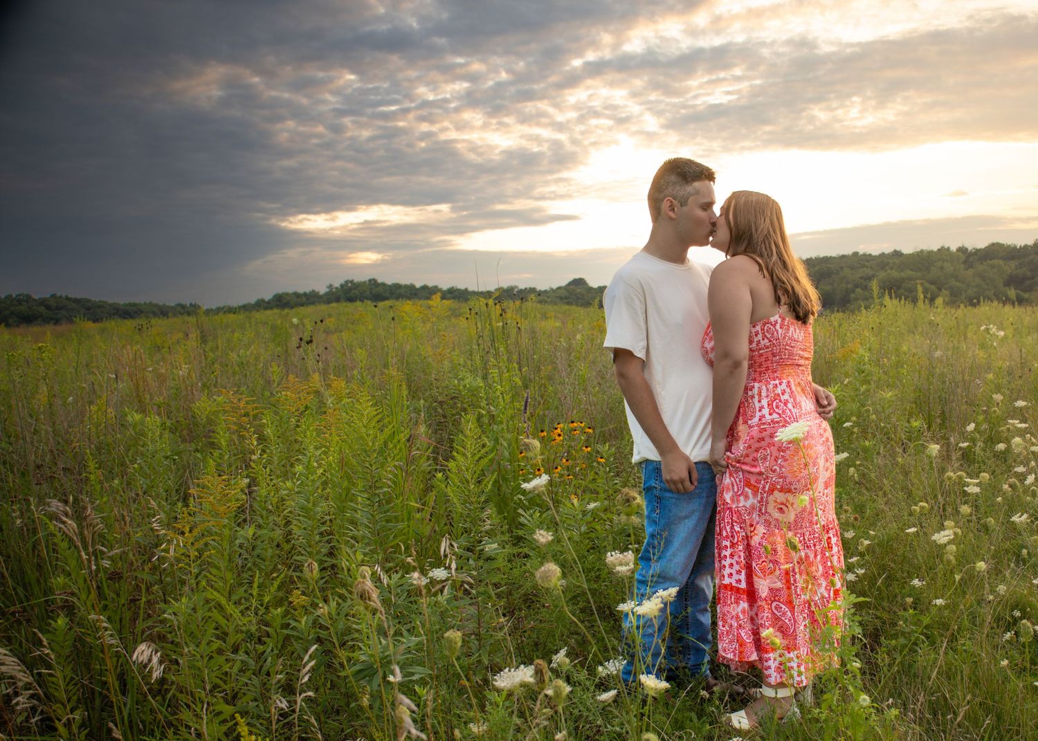 Young couple embracing in a wildflower field under dramatic cloudy sunset sky, sharing a tender romantic moment.