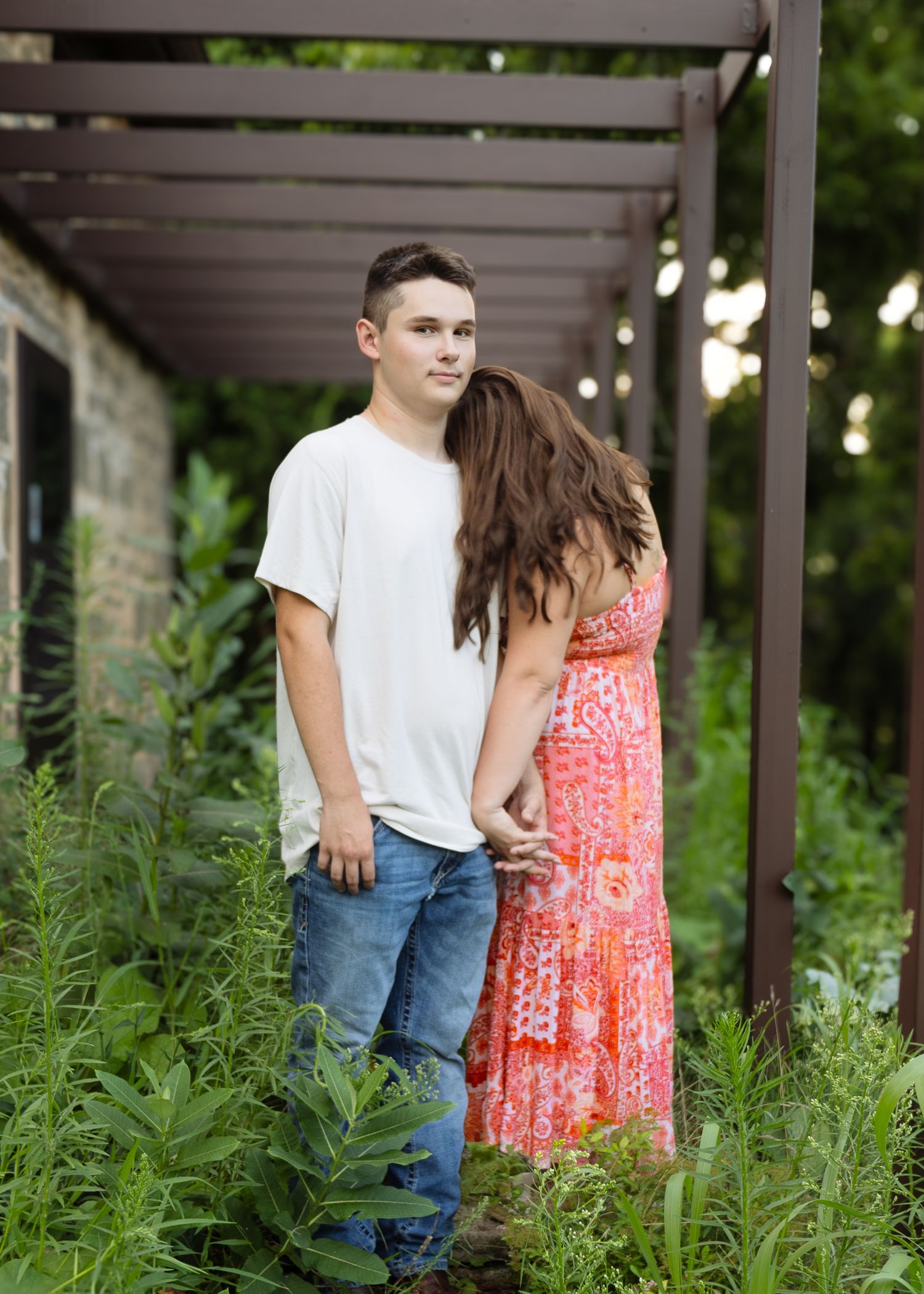 Young couple holding hands under a rustic pergola surrounded by lush green foliage outdoors.