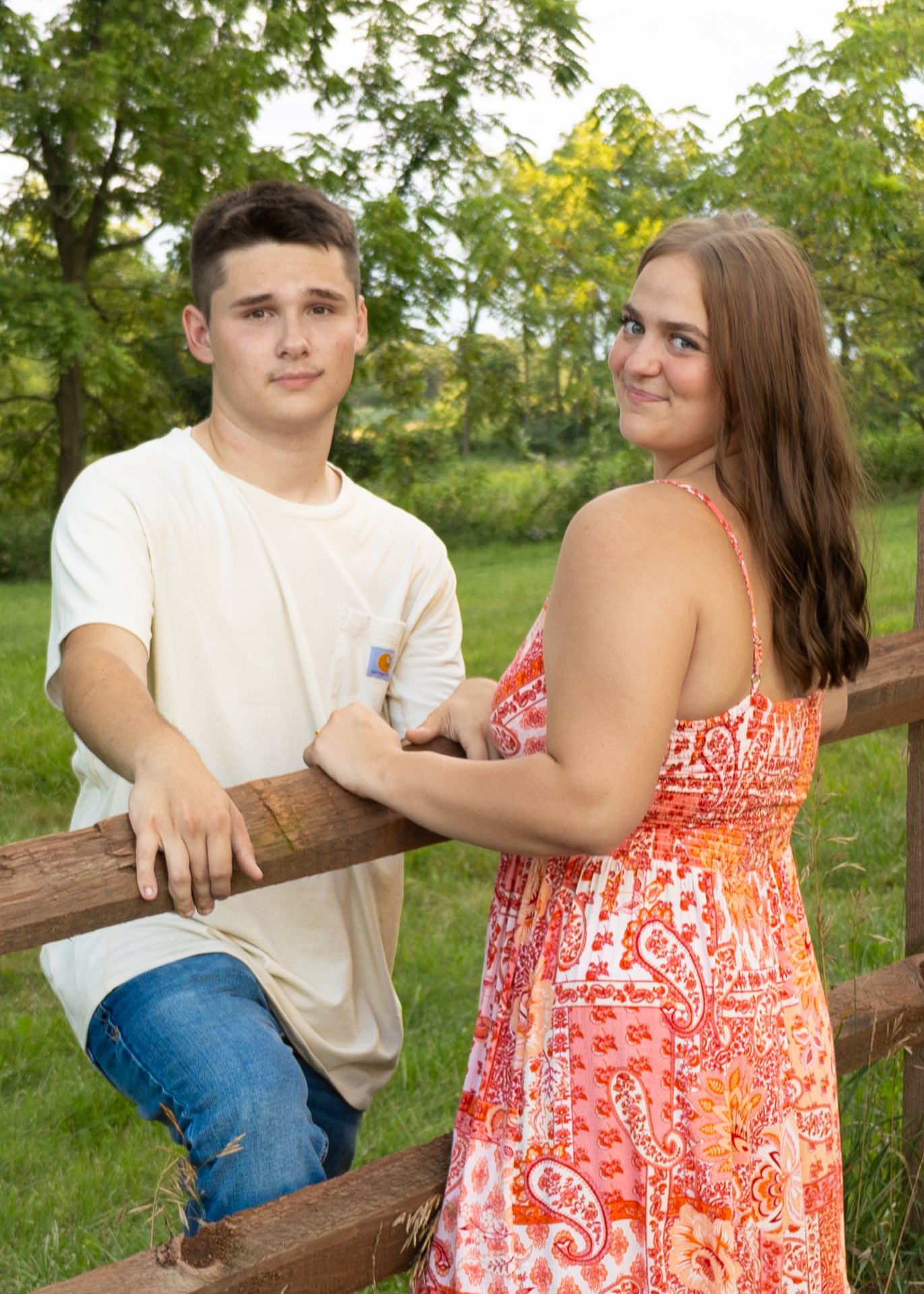 Young couple leaning on rustic wooden fence in green garden, smiling at camera on overcast day.