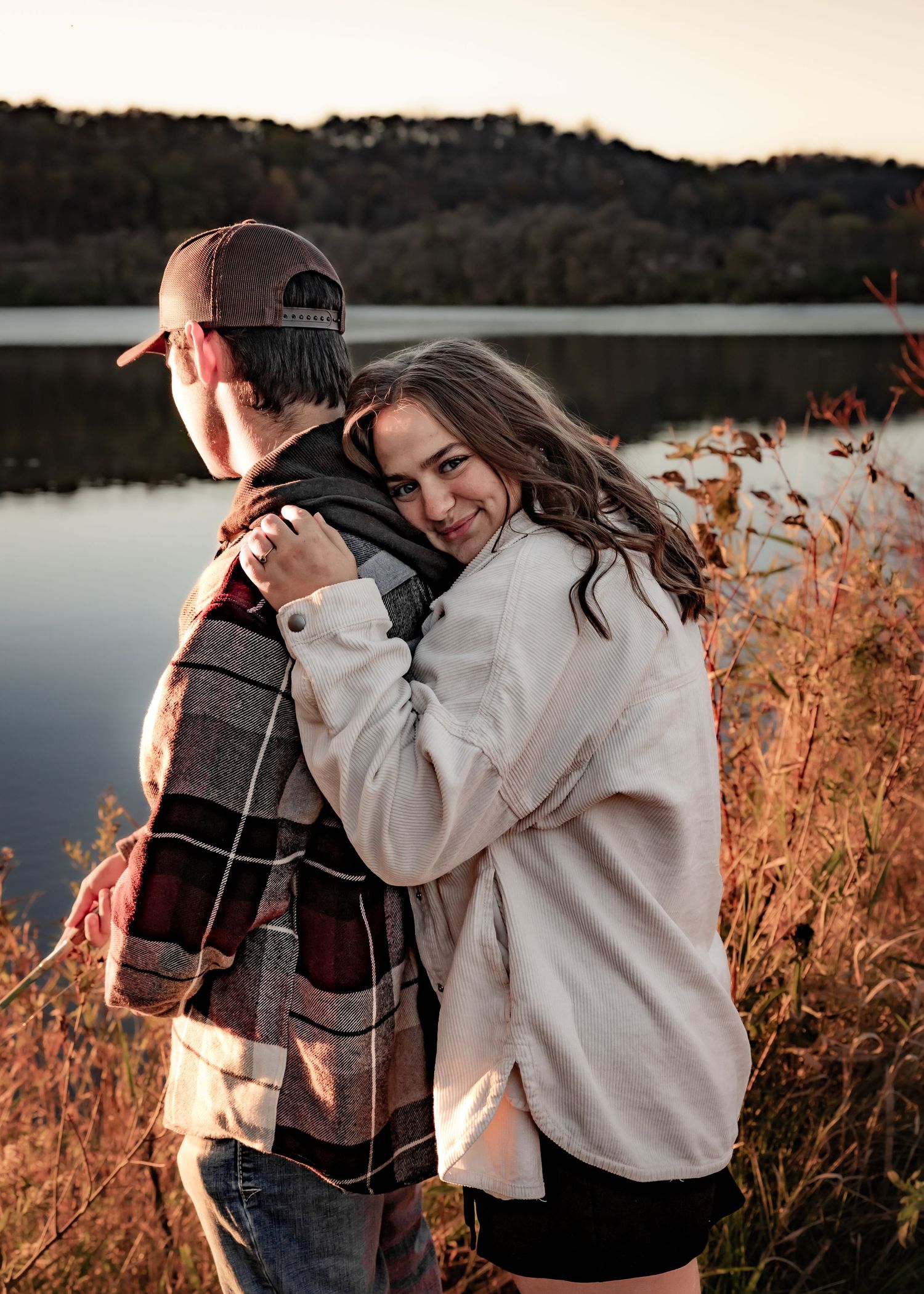 Woman hugging man from behind near a lake, smiling at camera during fall sunset.