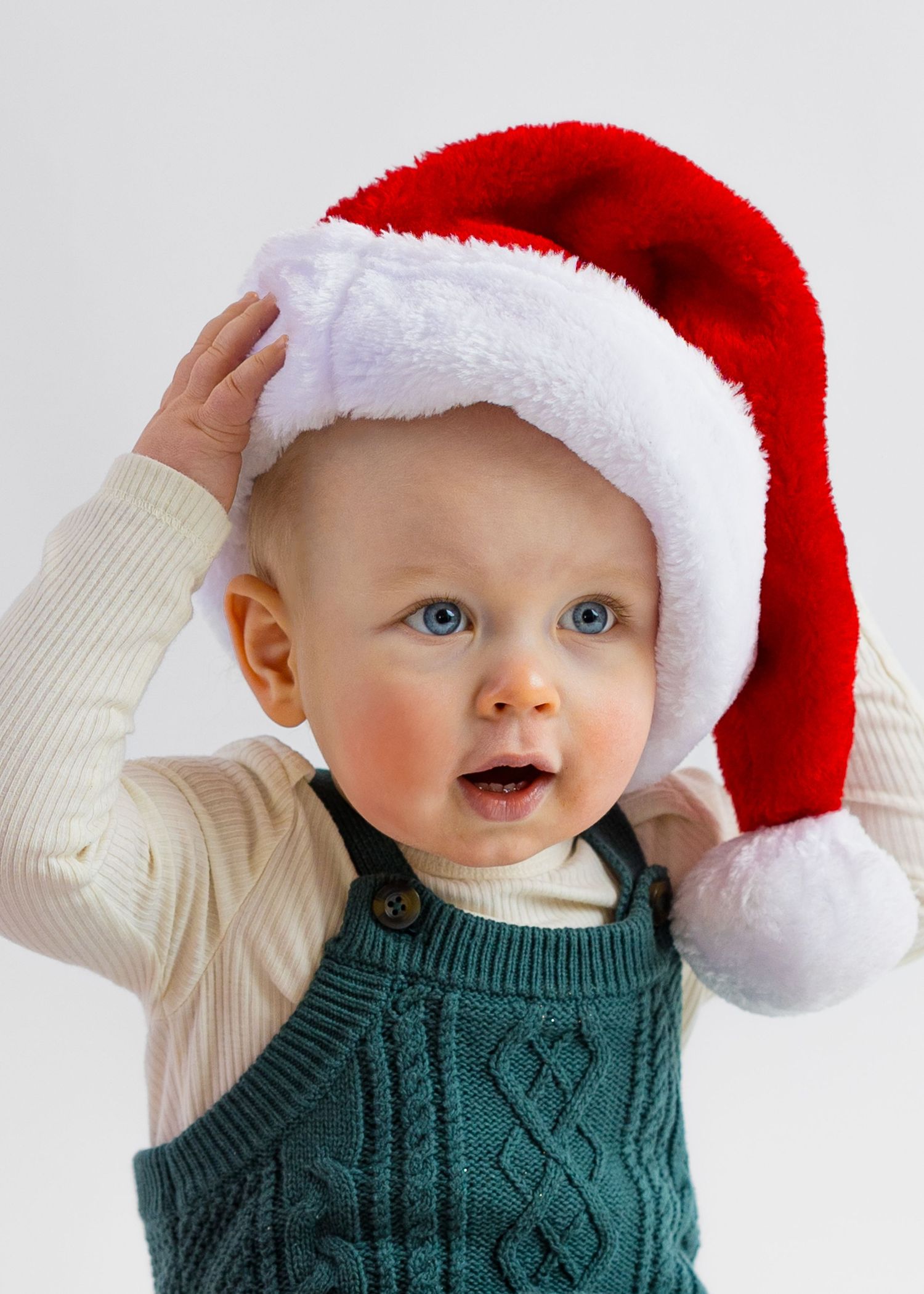 Blue-eyed baby in green knit overalls wearing an oversized red Santa hat looks up curiously.