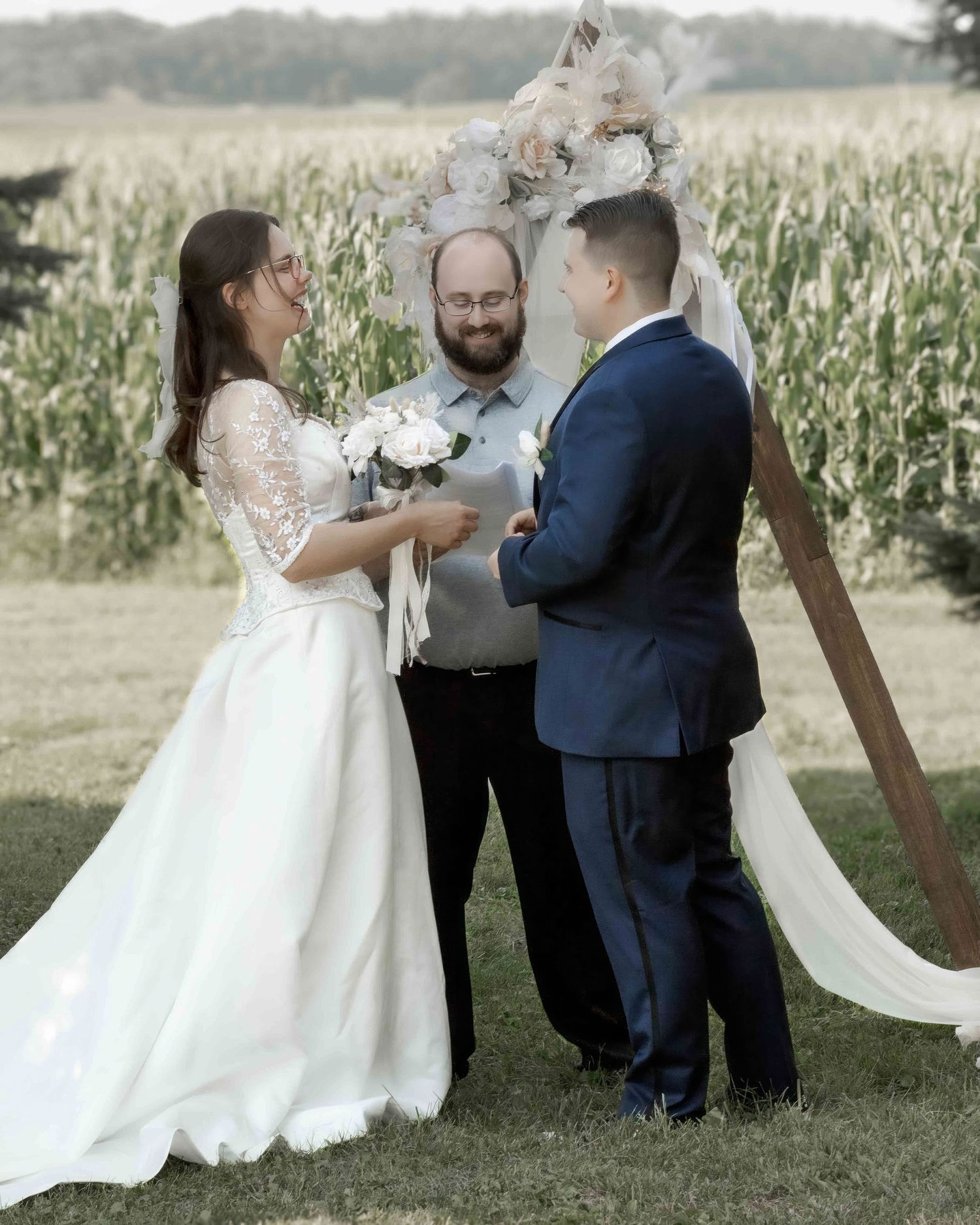 Bride and groom exchanging vows at rustic outdoor wedding ceremony near cornfield.