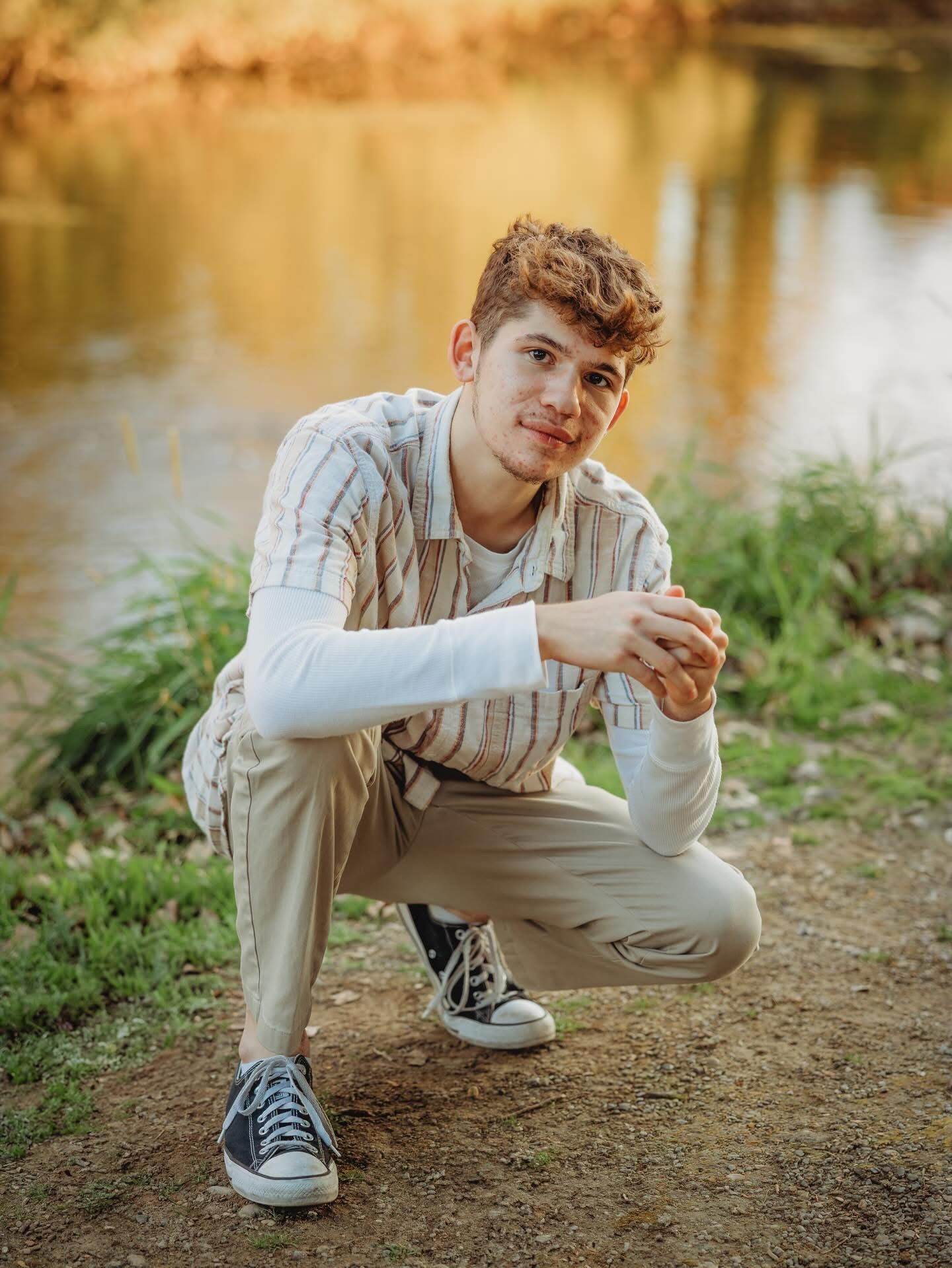 Young man in striped shirt crouching near a sunlit river bank for an outdoor senior portrait.