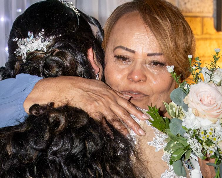 Emotional woman with tears in her eyes hugs a bride wearing a floral hairpiece while holding a bouquet of pink roses at a wedding.