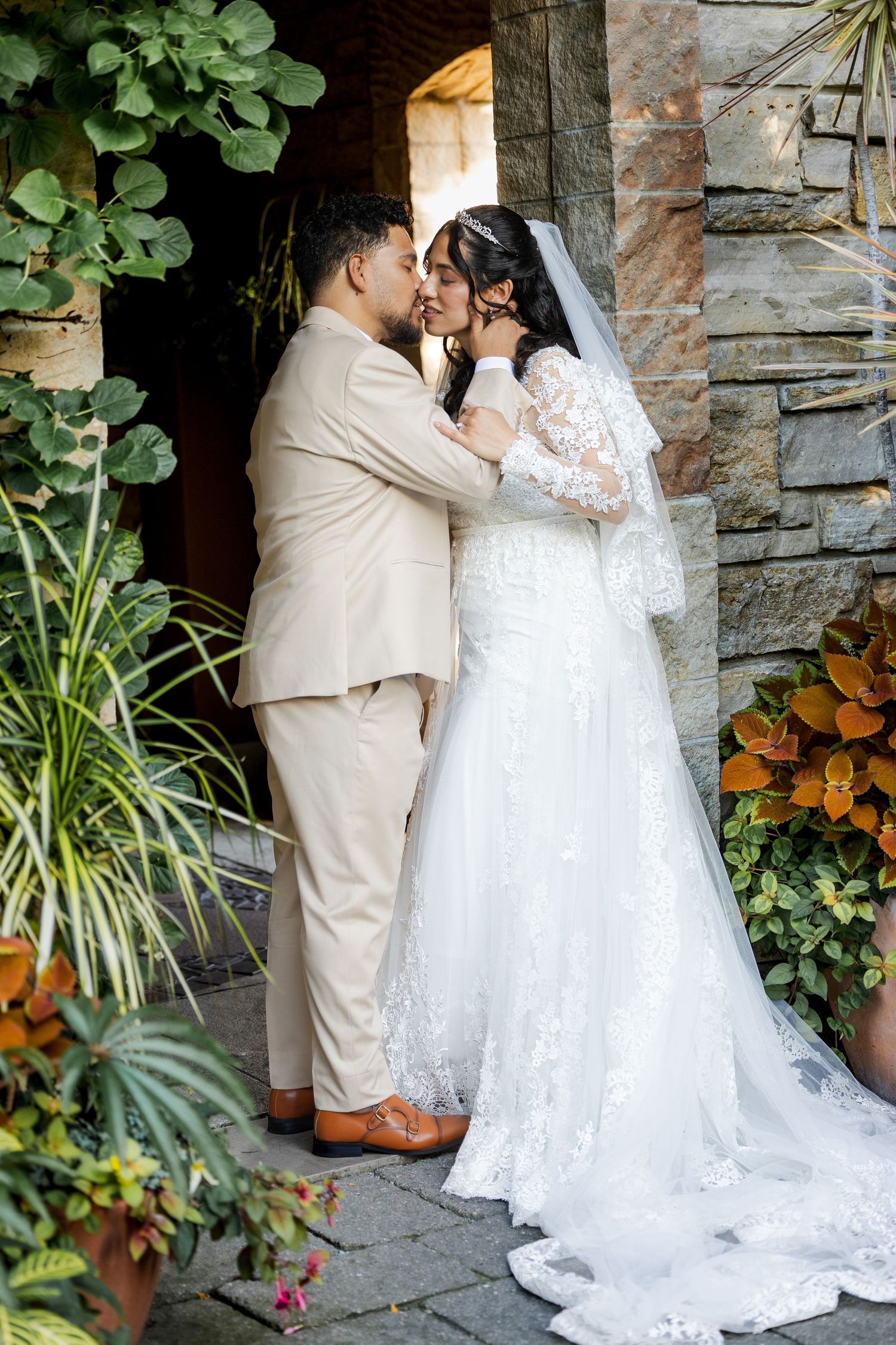 Bride in a lace gown and veil kisses groom in a tan suit beside a stone wall and lush green plants on a wedding day.