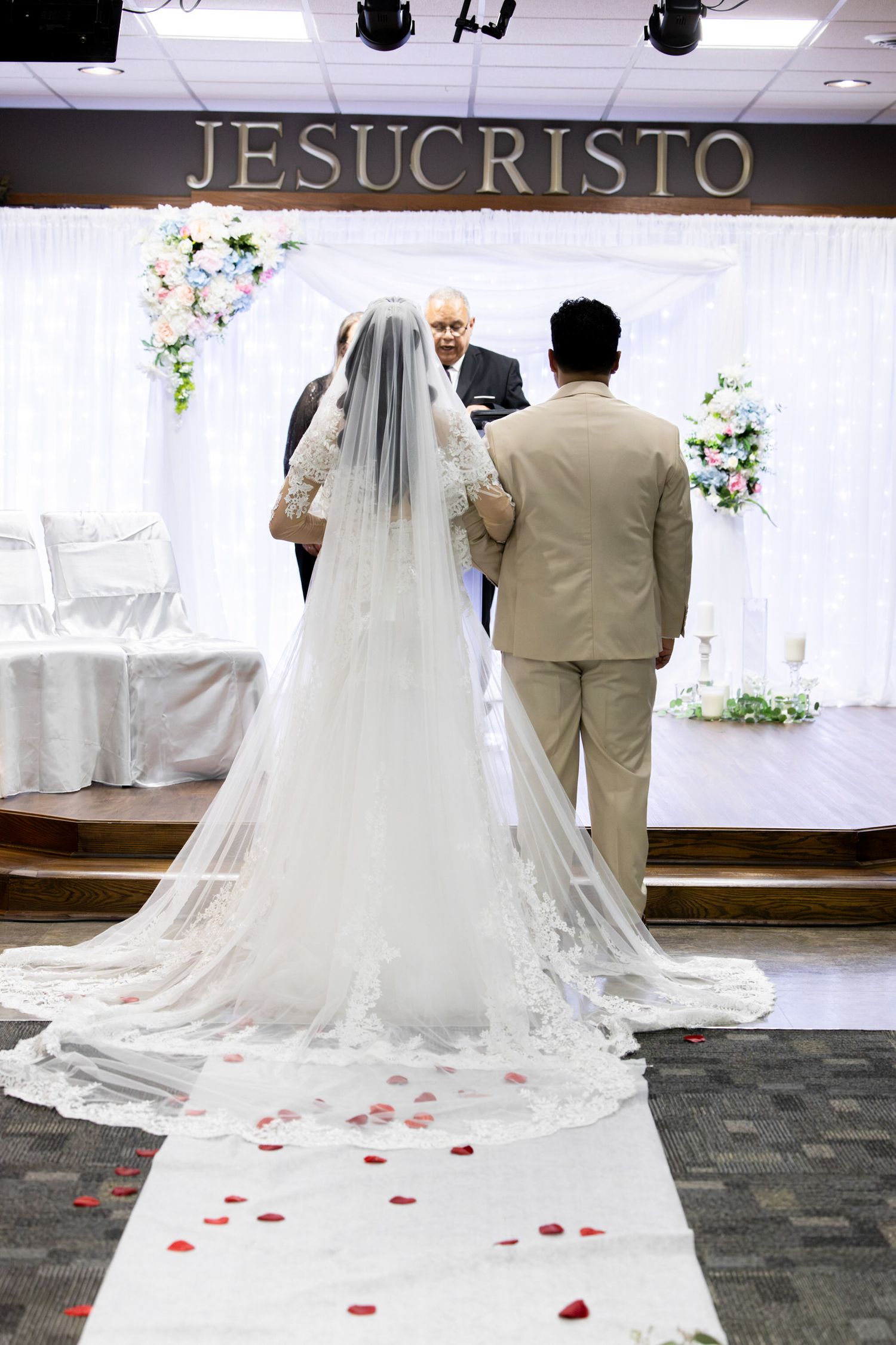 Bride in a long lace veil and groom in a tan suit stand at the altar during a wedding ceremony, with rose petals lining the white aisle.