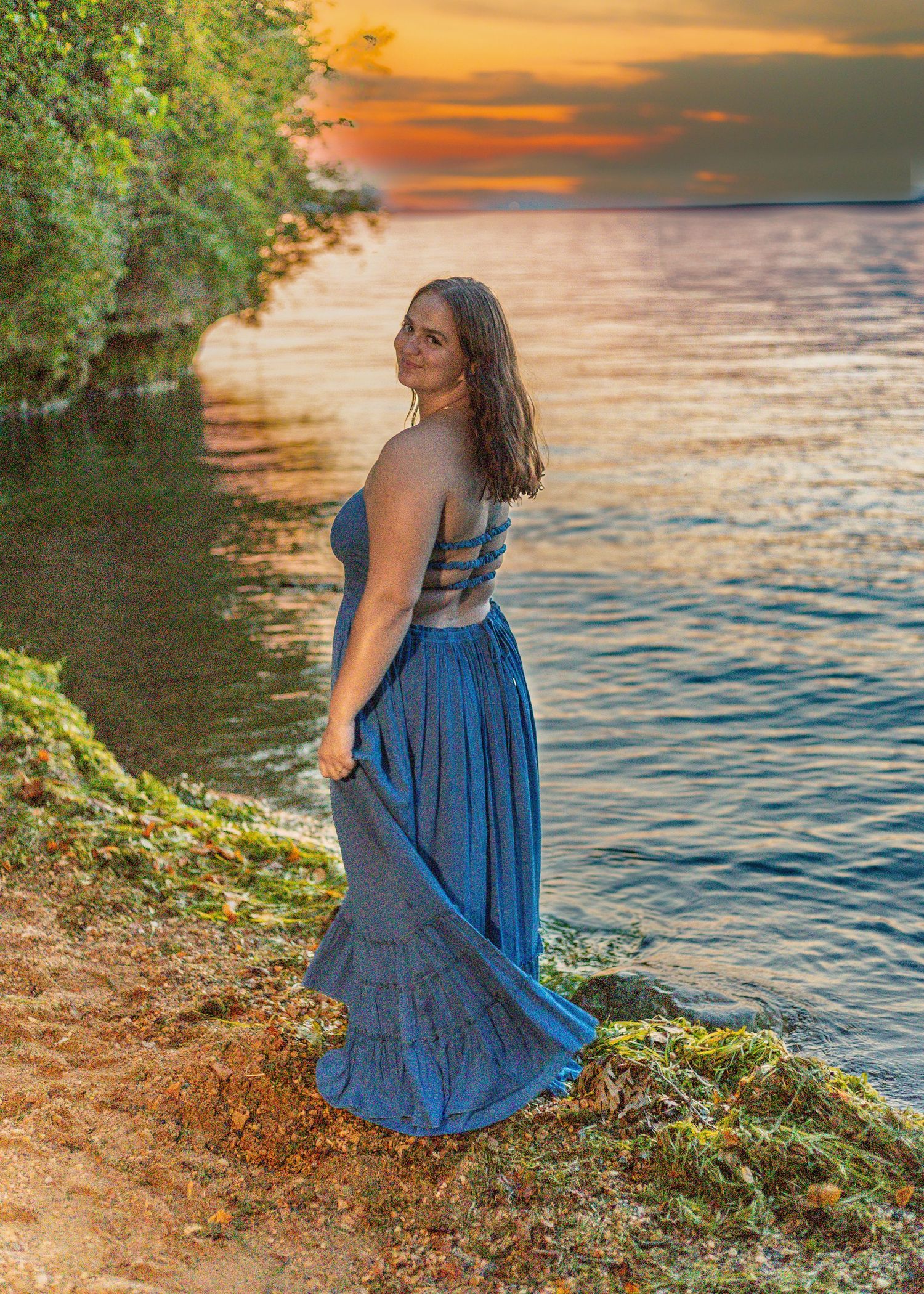 Woman in blue maxi dress poses by waterfront at sunset, rocky shore with lush greenery backdrop.