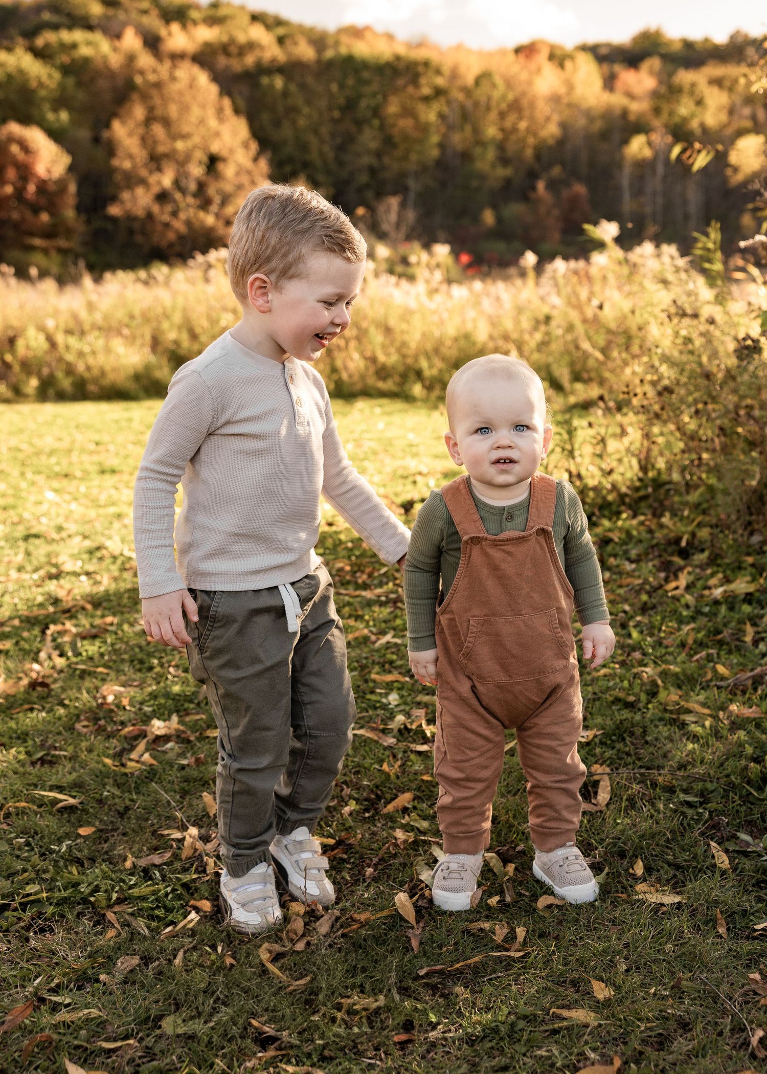Two young brothers standing together in a sunlit autumn field, older sibling smiling at baby.