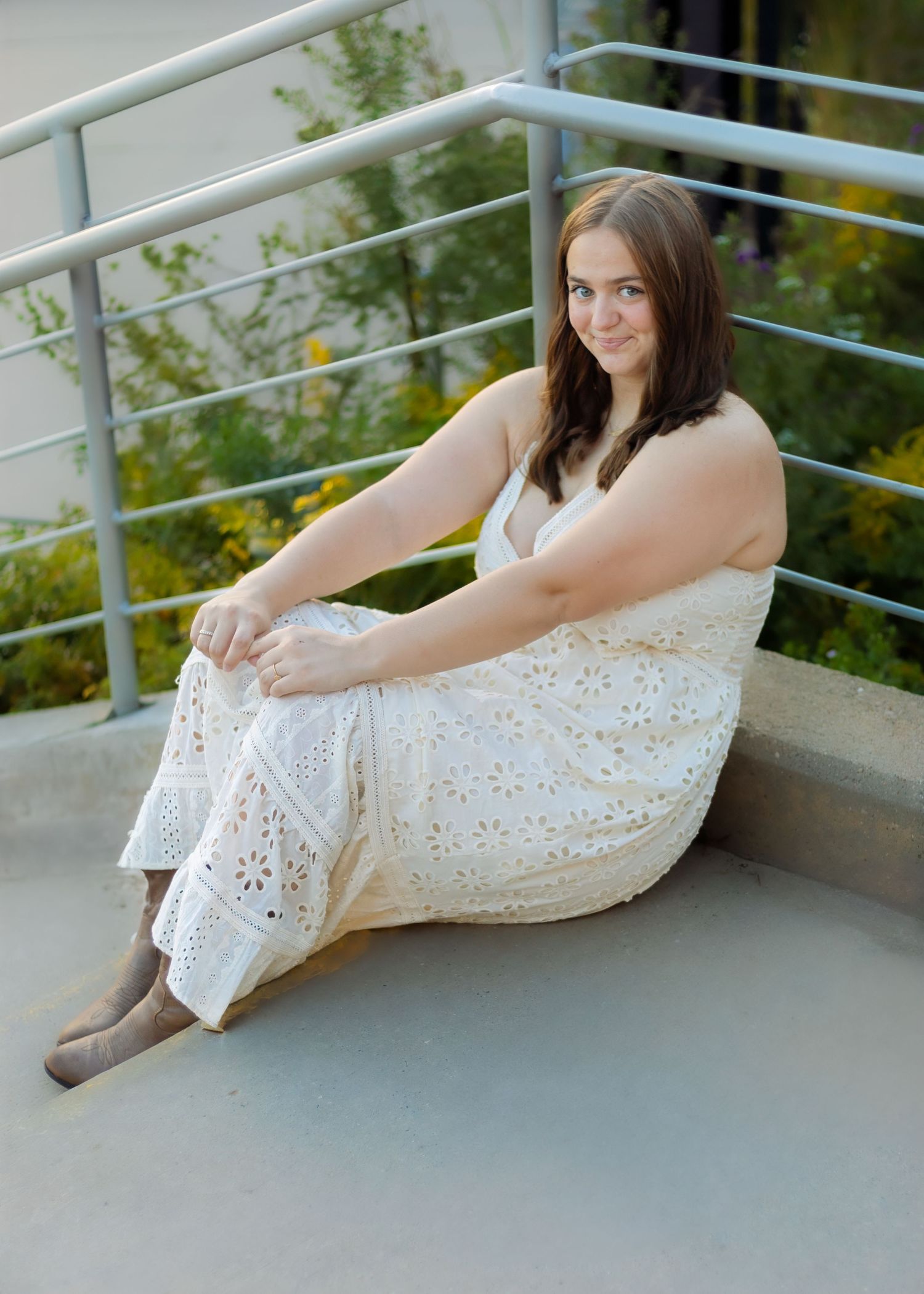 A young woman in a white eyelet maxi dress sits on outdoor steps smiling at the camera.