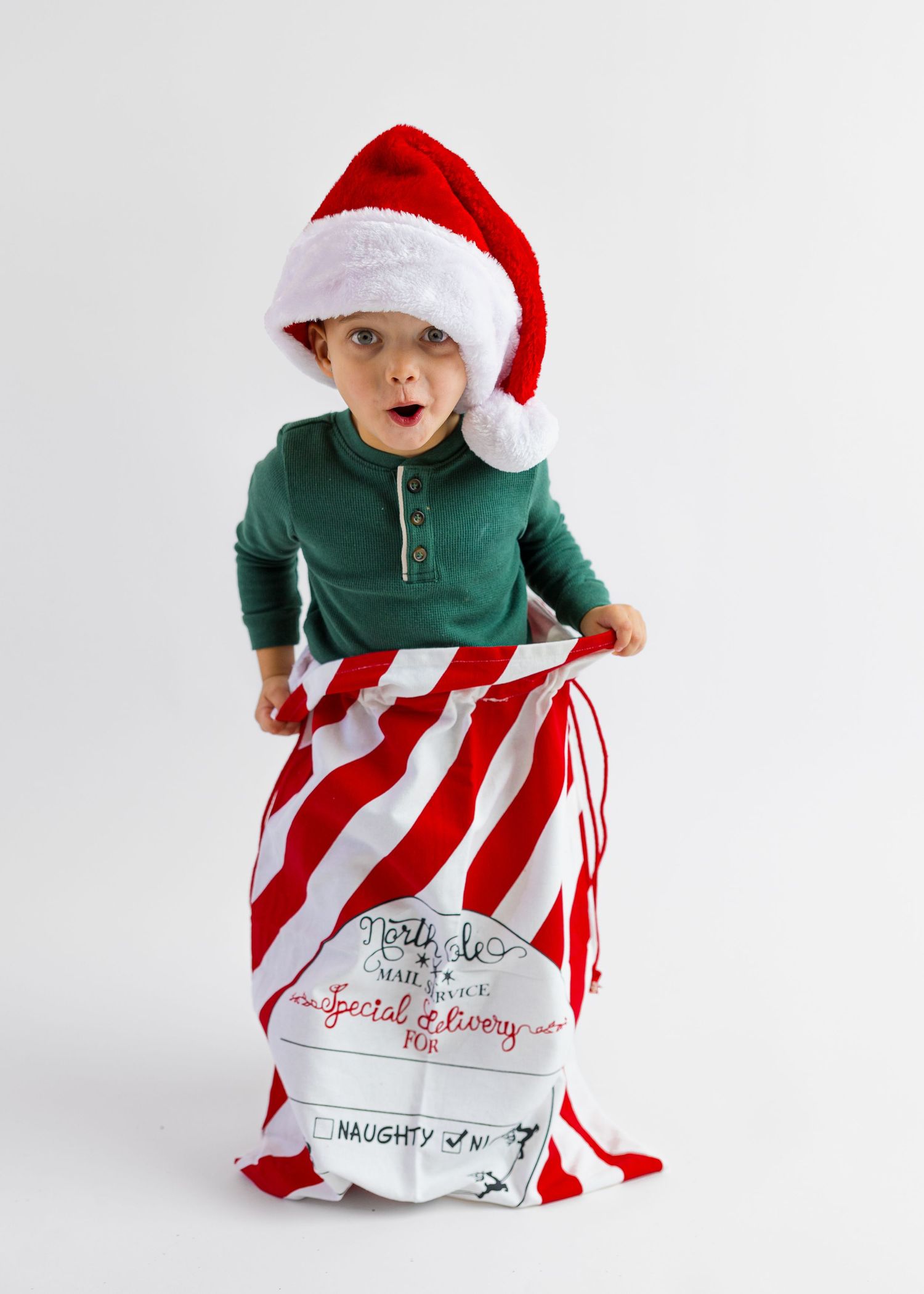 Surprised toddler in green shirt and Santa hat stands inside a large red striped Christmas bag.