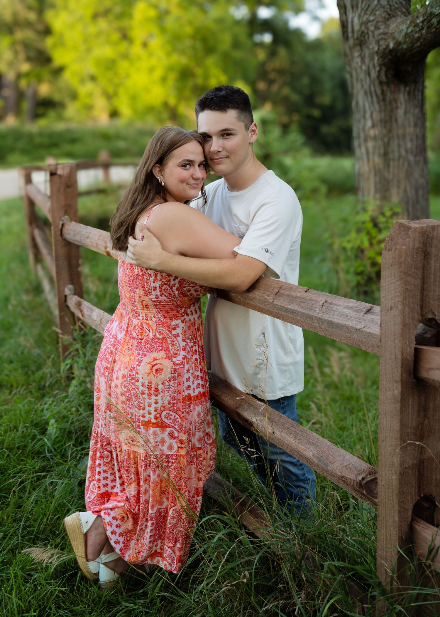 Young couple embracing beside a rustic wooden fence surrounded by lush greenery.