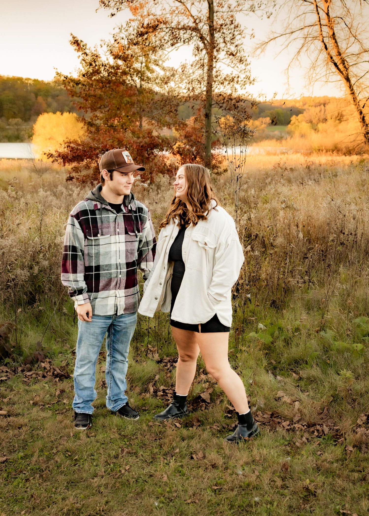 Young couple holding hands in autumn field at sunset wearing casual flannel and white jacket.