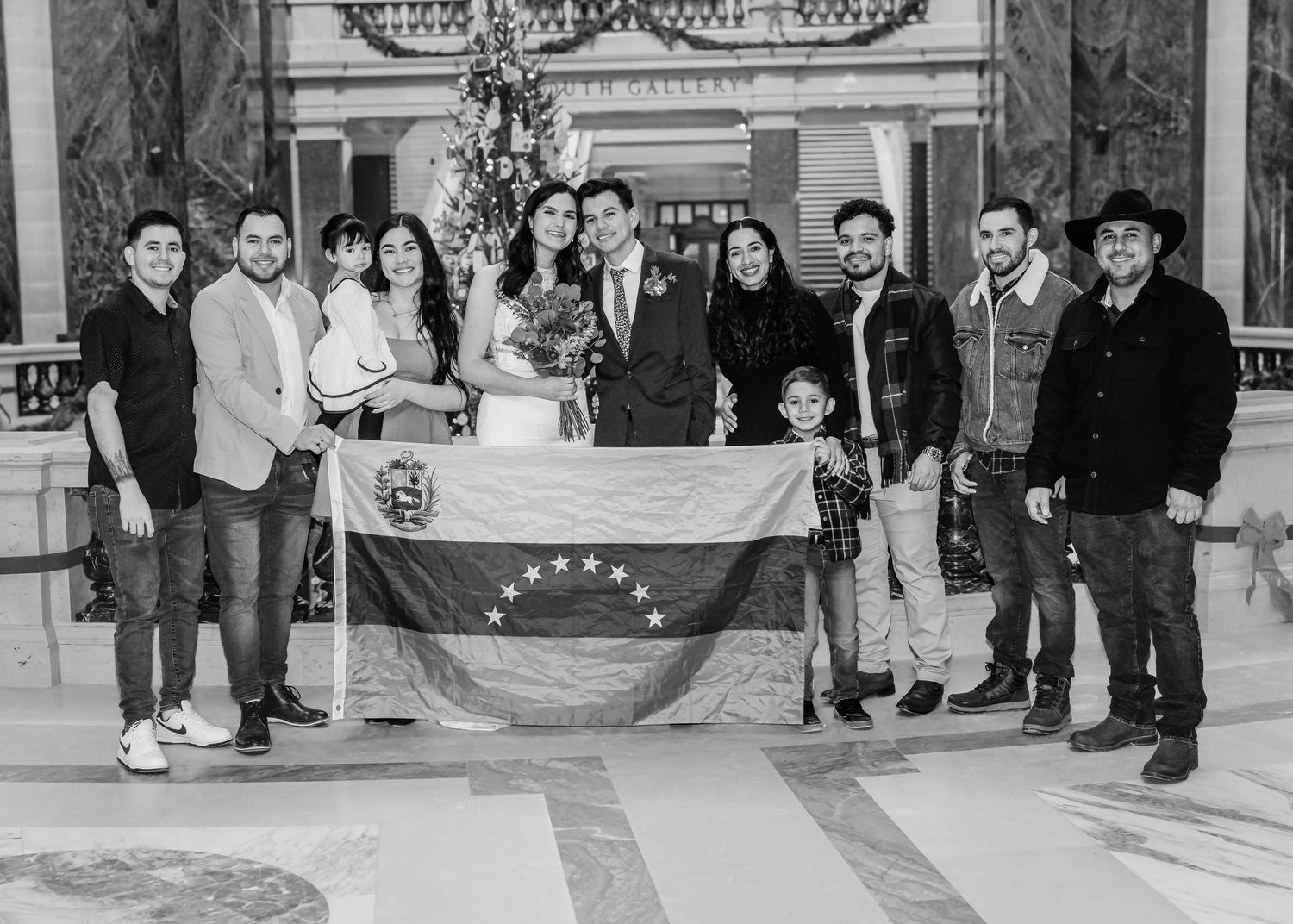 Black and white wedding group photo with family holding a Venezuelan flag in a decorated hall.