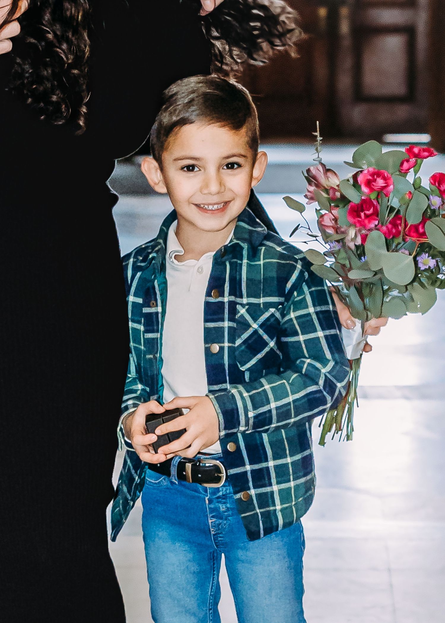 Adorable young boy in plaid shirt smiling while holding a bouquet of pink flowers at a wedding.