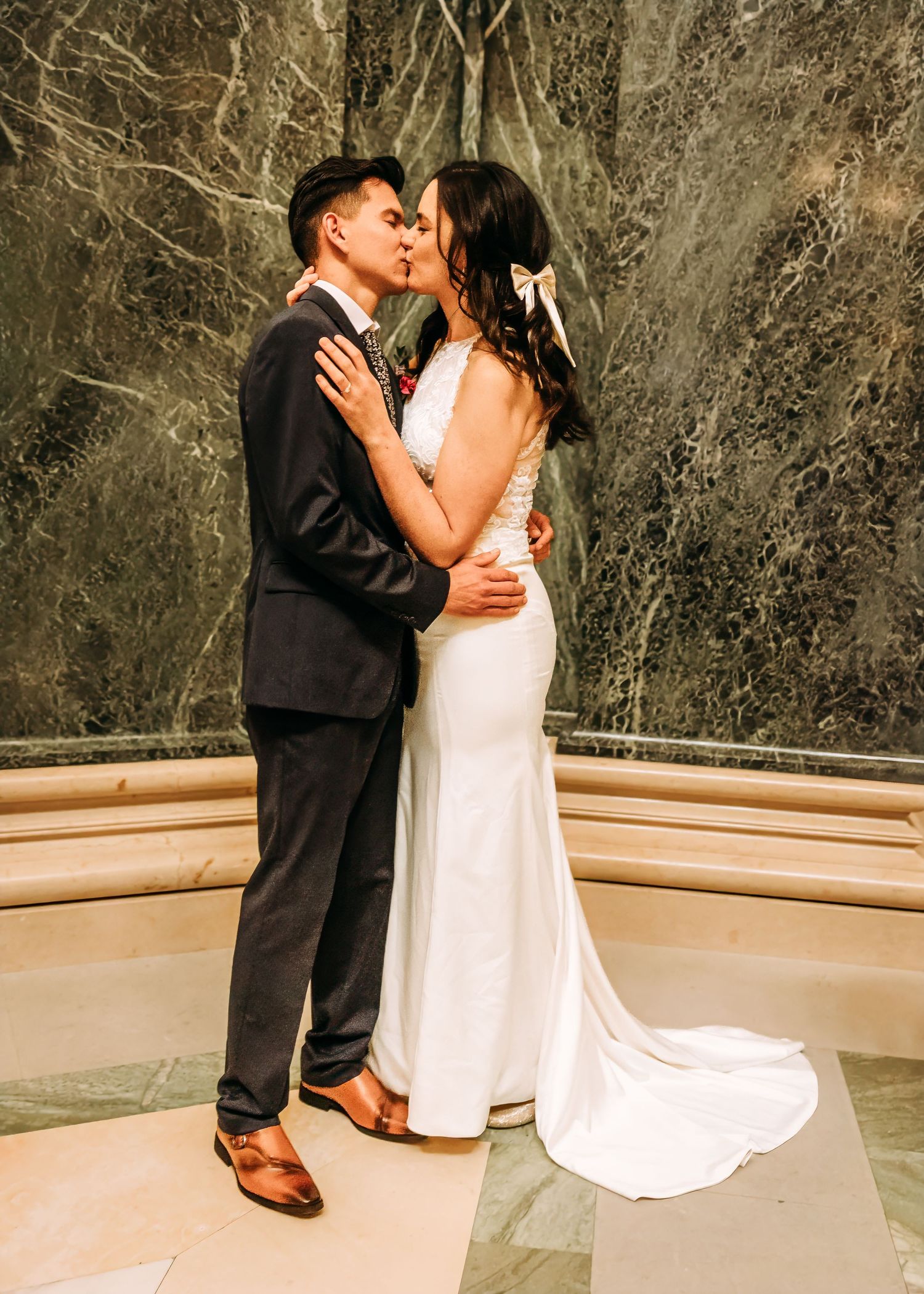 Newlyweds share a romantic kiss, bride in fitted white gown with bow, groom in dark suit.