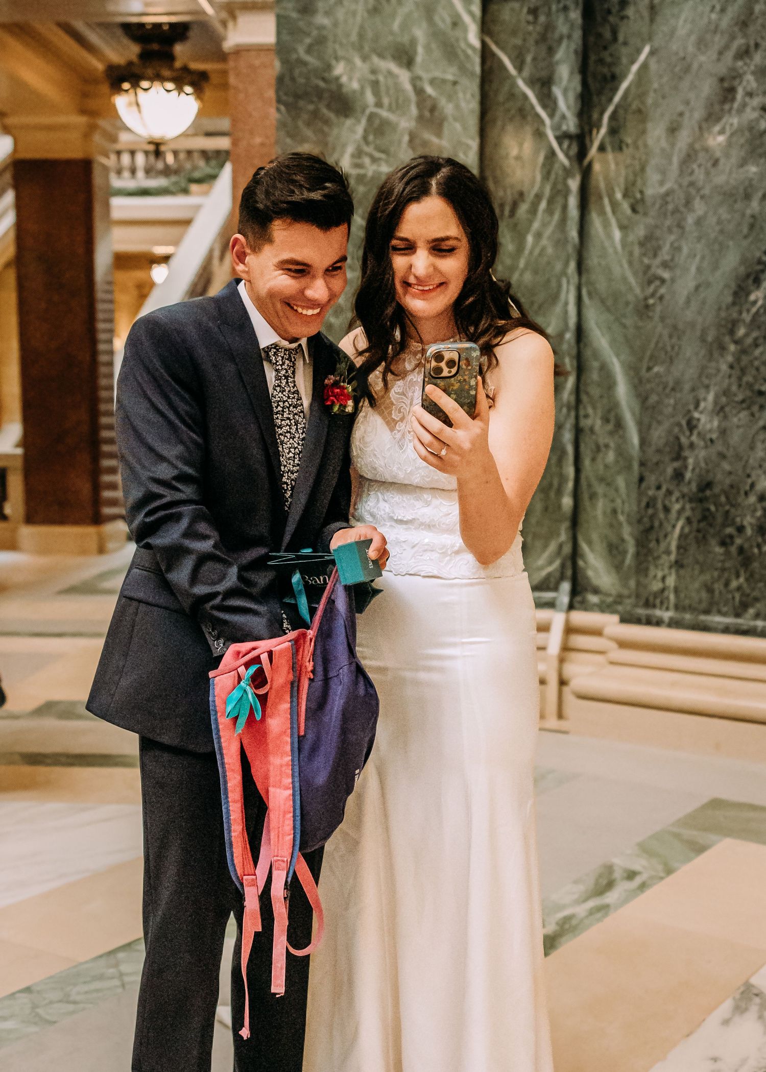 Joyful bride and groom smiling at phone screen, groom holding colorful ribbon wands indoors.