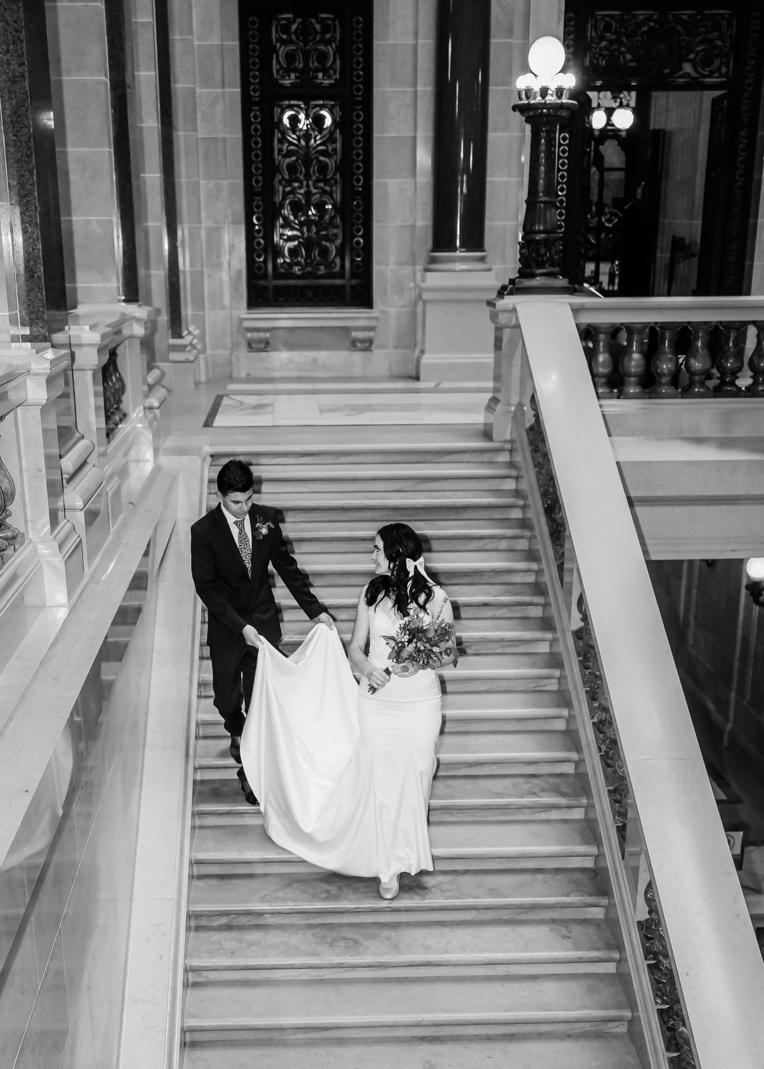Black and white photo of bride and groom descending ornate marble staircase in historic building.