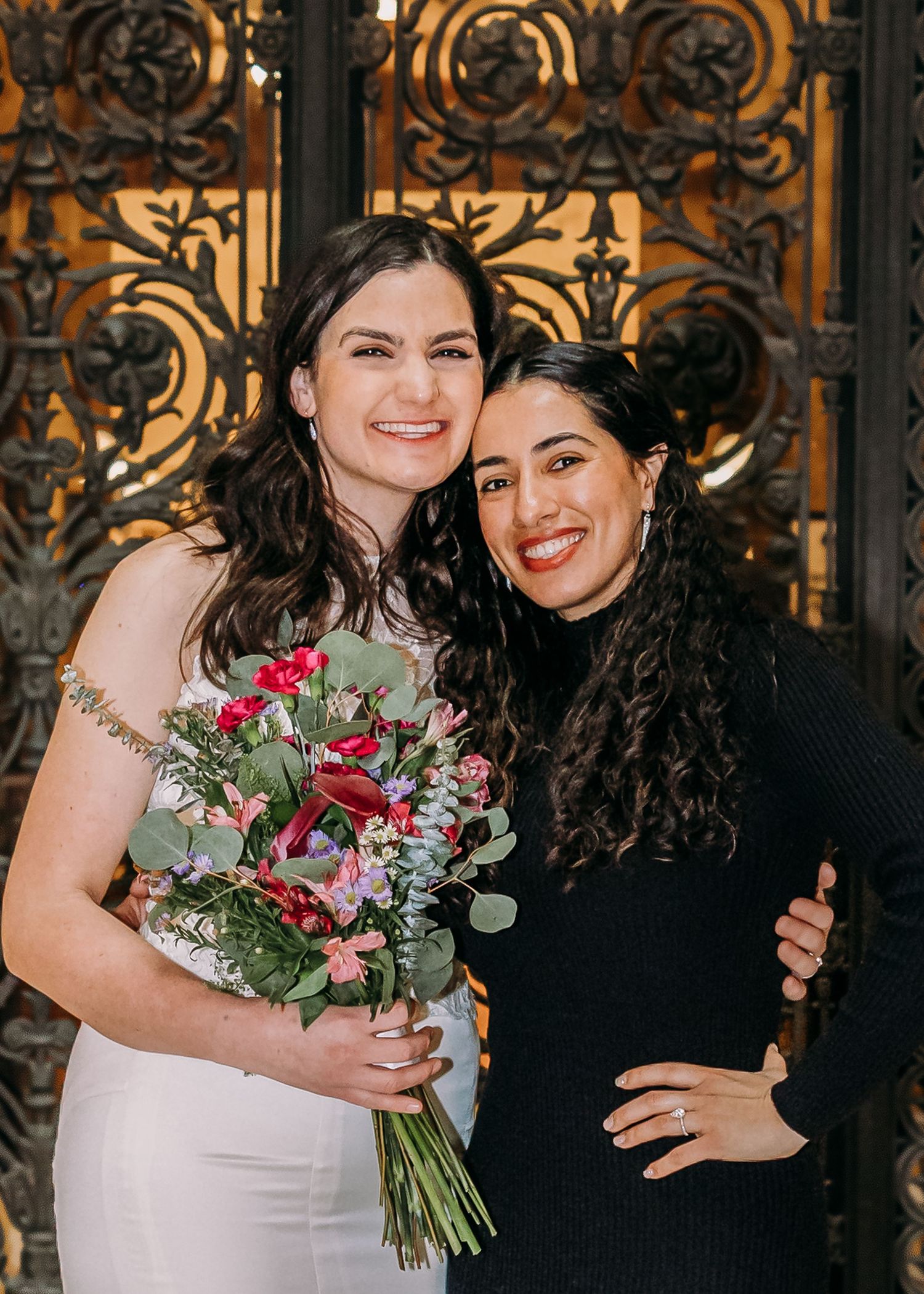 Smiling bride holding colorful bouquet poses with dark-haired guest in black sweater before decorative iron gates.