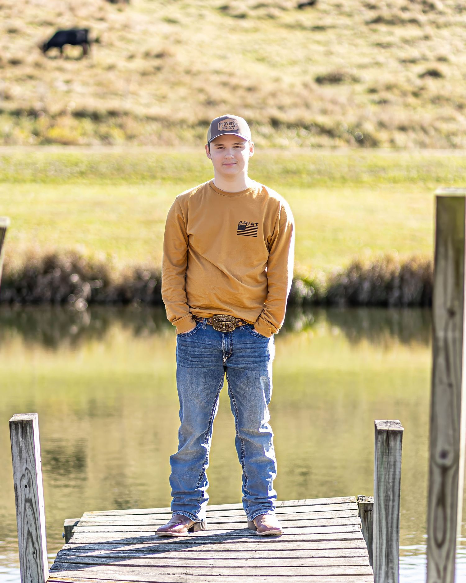 Young man in mustard sweatshirt and jeans stands on a wooden dock by a calm pond on a sunny day.