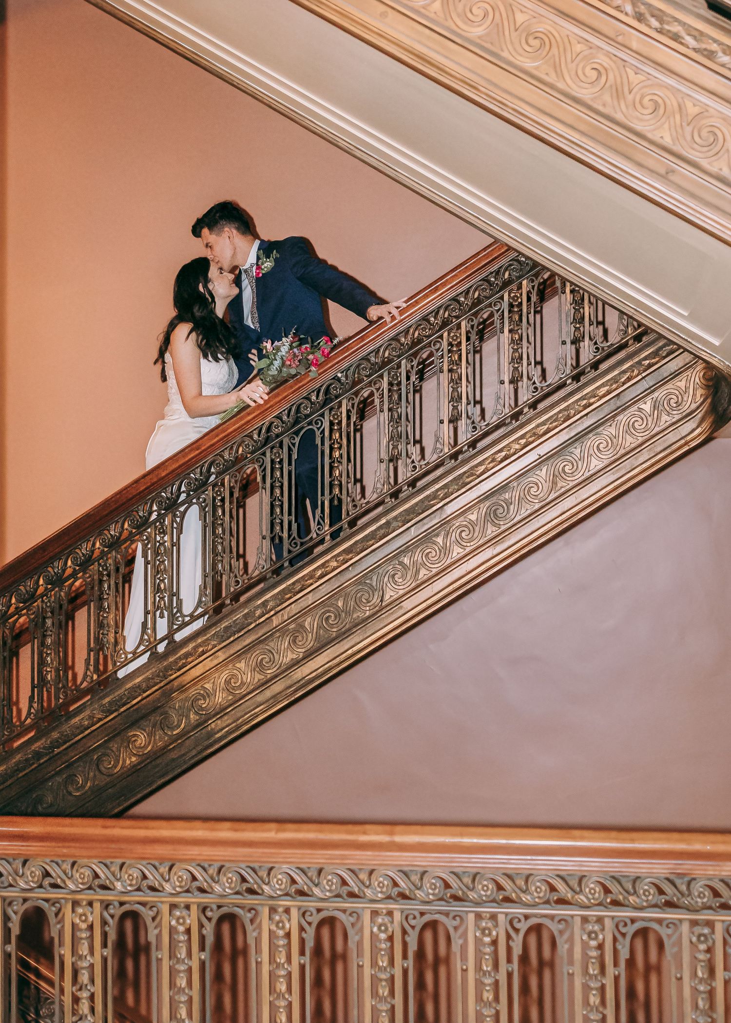 Bride and groom sharing a romantic kiss on an ornate golden staircase railing at a elegant wedding venue.