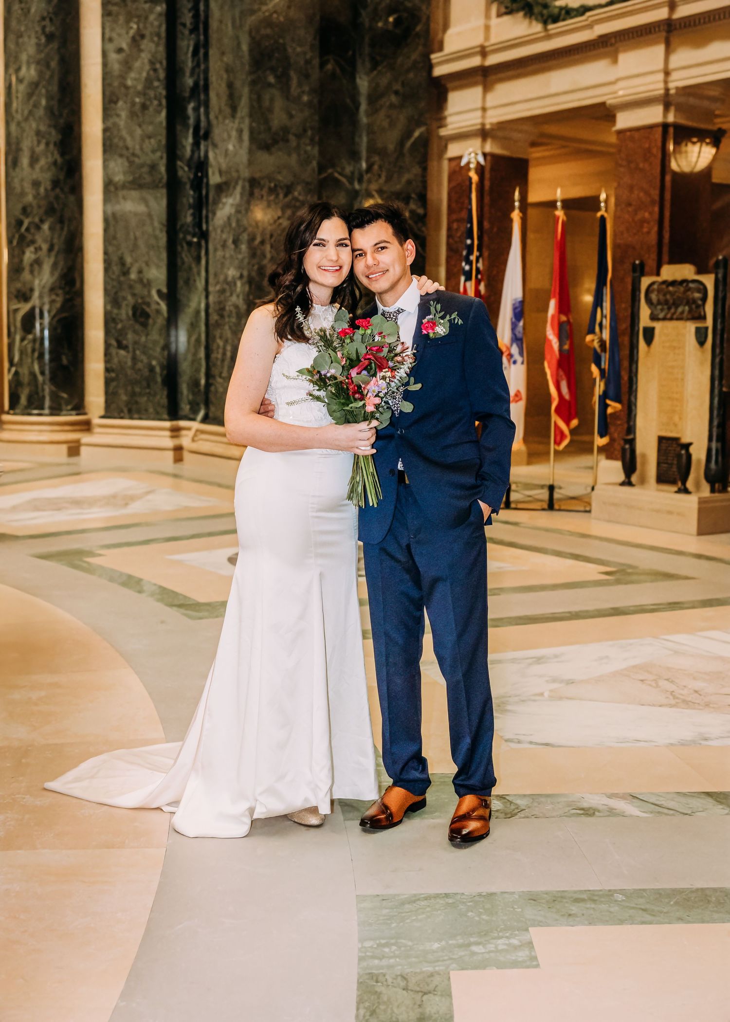 Smiling newlyweds pose in marble hall, bride in white gown with bouquet and groom in navy suit.