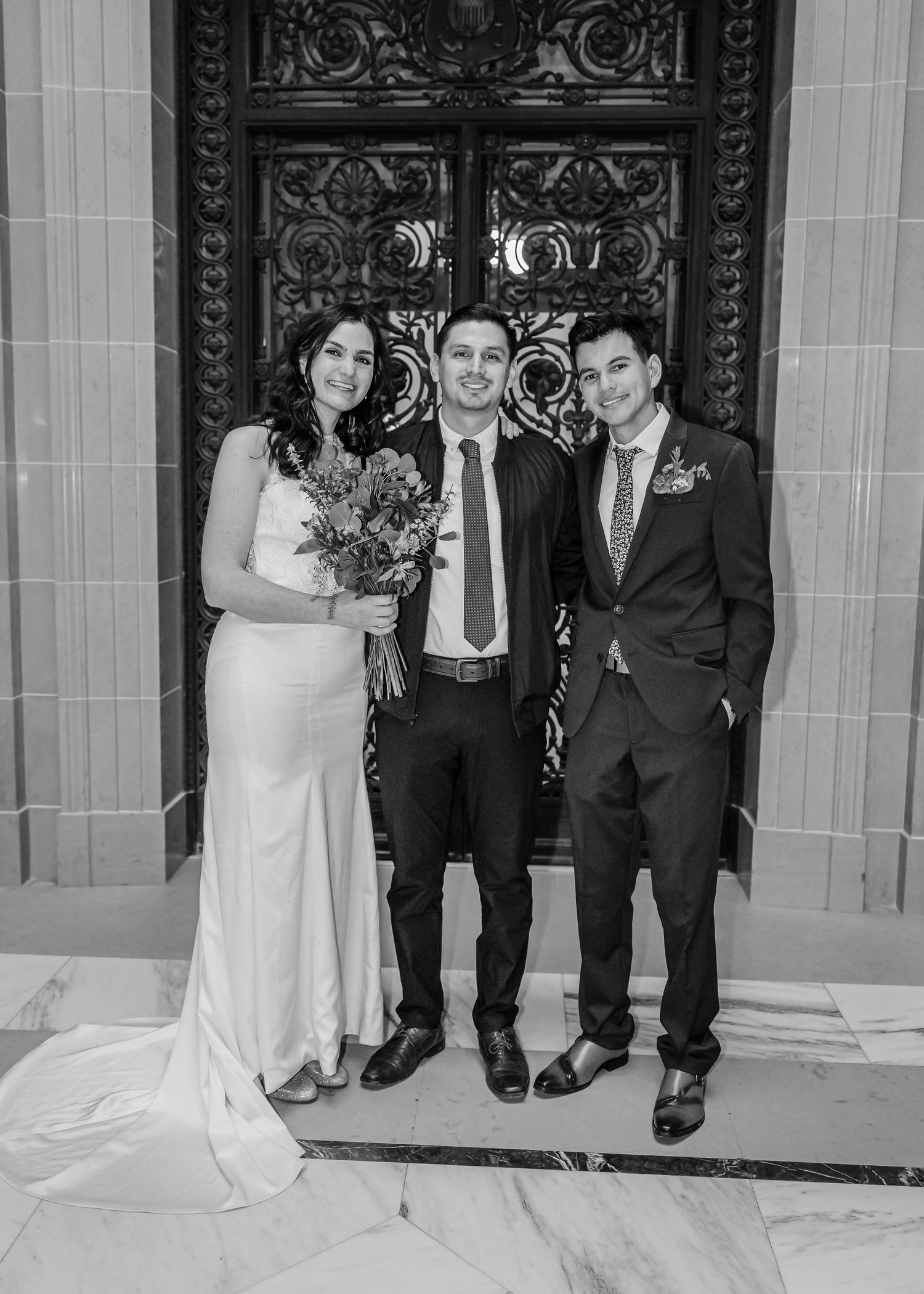 A bride and groom pose with a guest in front of ornate doors in a black and white wedding photo.