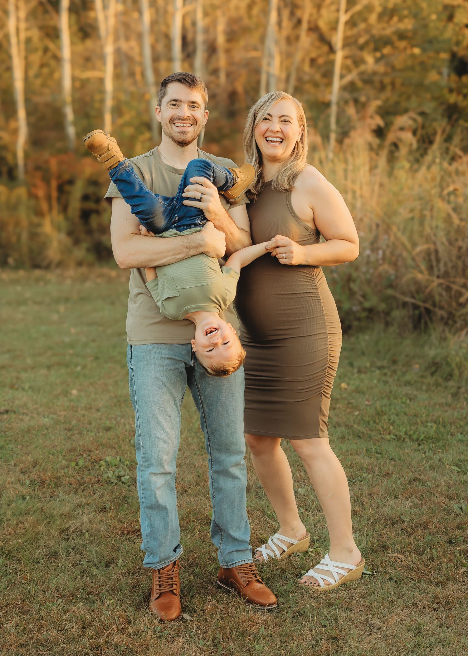 A smiling family poses outdoors as the father playfully holds their toddler upside down.
