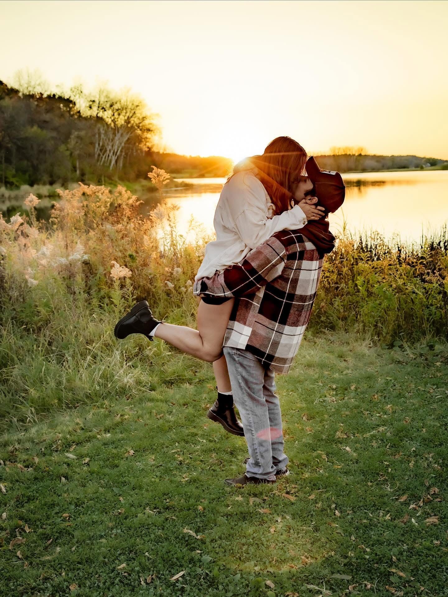 Couple sharing a romantic kiss at golden sunset by a lake, man lifting woman in plaid blanket wrap.