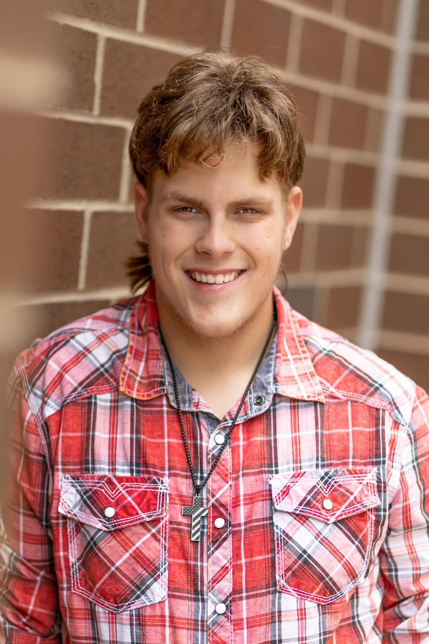 Smiling teenage boy in red plaid western shirt poses casually against brick wall outdoors.