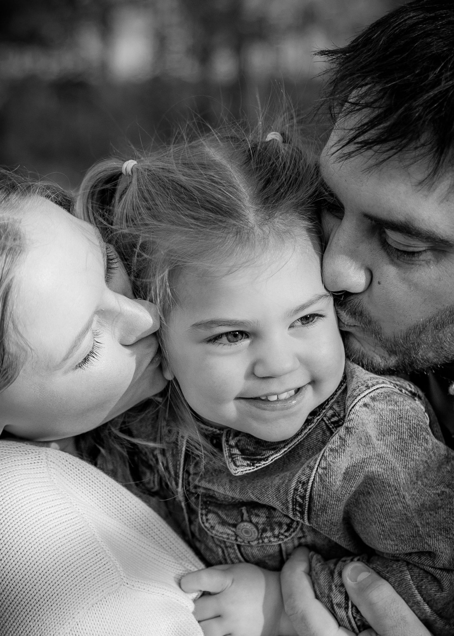 Black and white photo of parents kissing their smiling young daughter on both cheeks.