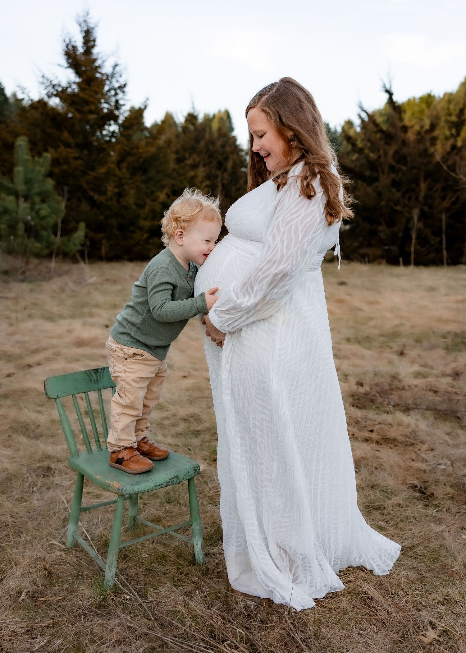 A toddler boy stands on a green chair kissing a pregnant womans belly in an outdoor field.