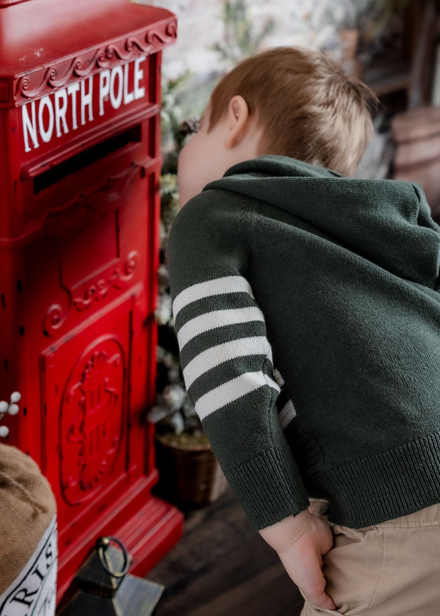 Young boy in green striped hoodie peering into red North Pole mailbox during Christmas mini session.