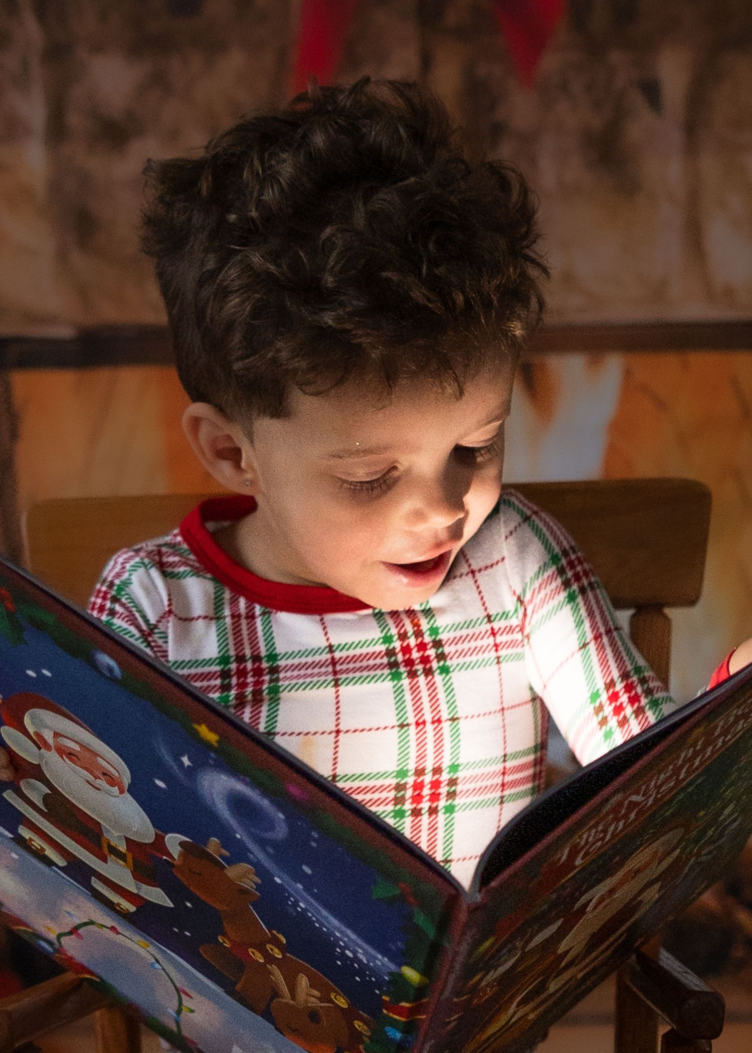Curly-haired boy in plaid Christmas pajamas reading a glowing holiday book with wonder.