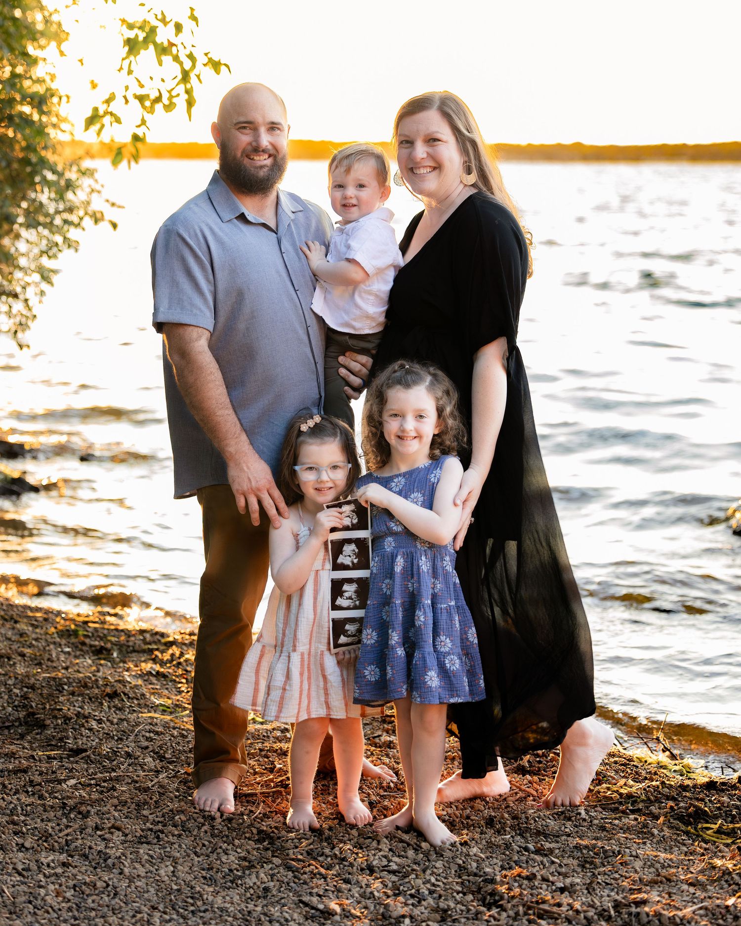 Family of five poses barefoot by a lake at golden hour, with two girls holding an ultrasound photo announcing a new baby.