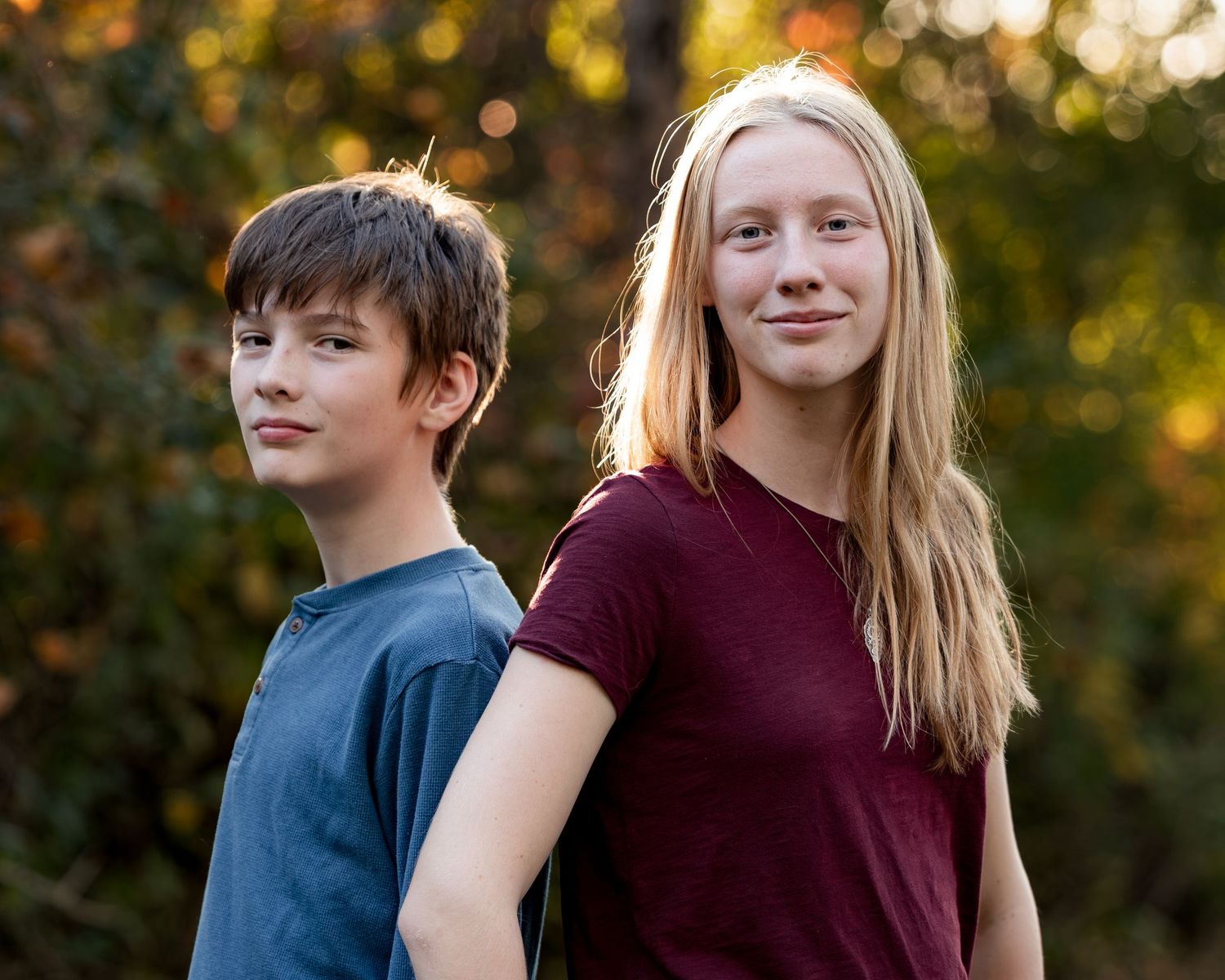 Two teenagers stand back to back outdoors in golden sunlight, a boy in a blue shirt and a girl with long blonde hair in a maroon shirt.