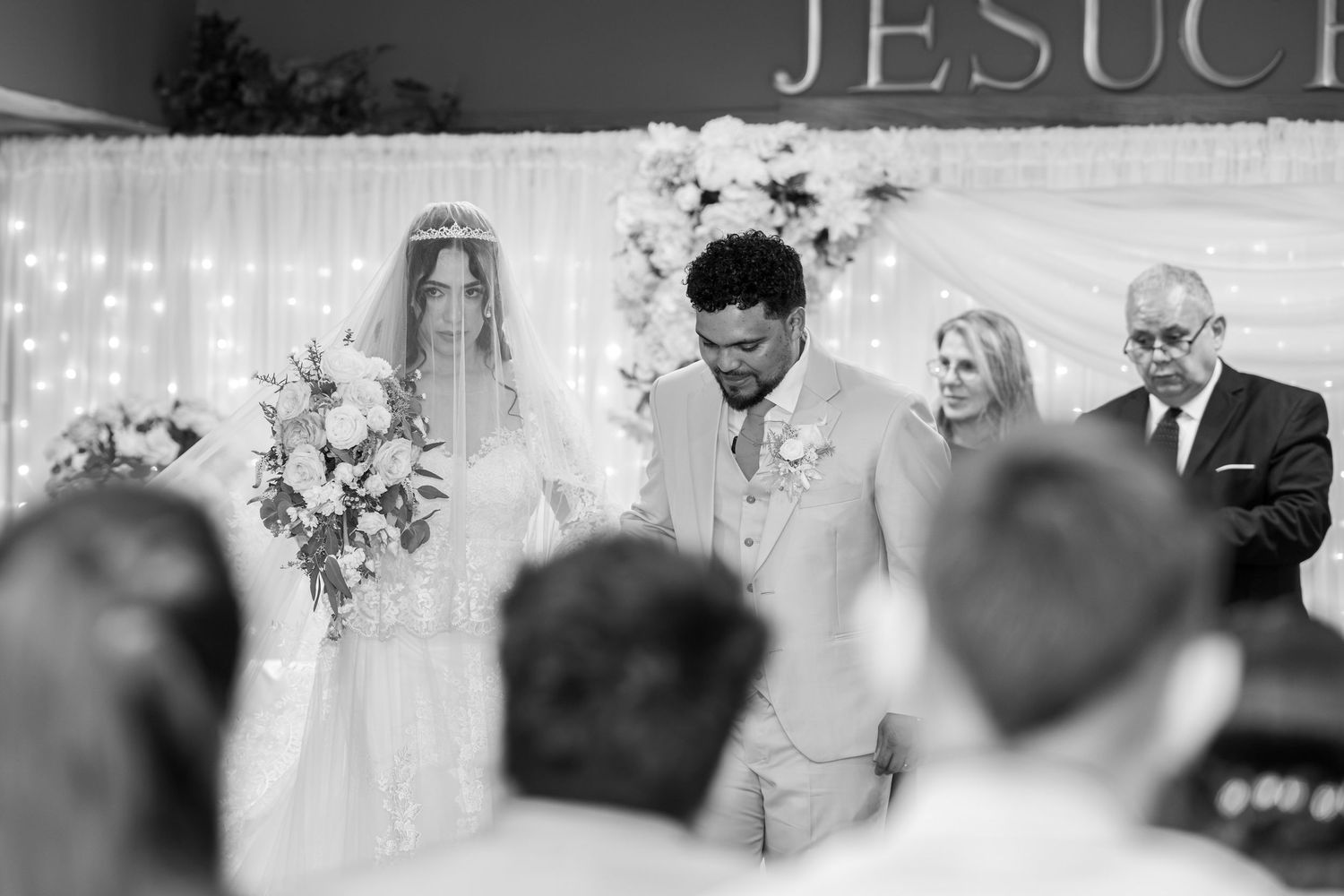 Black and white wedding ceremony with a veiled bride holding a floral bouquet beside a groom in a light suit as guests look on.