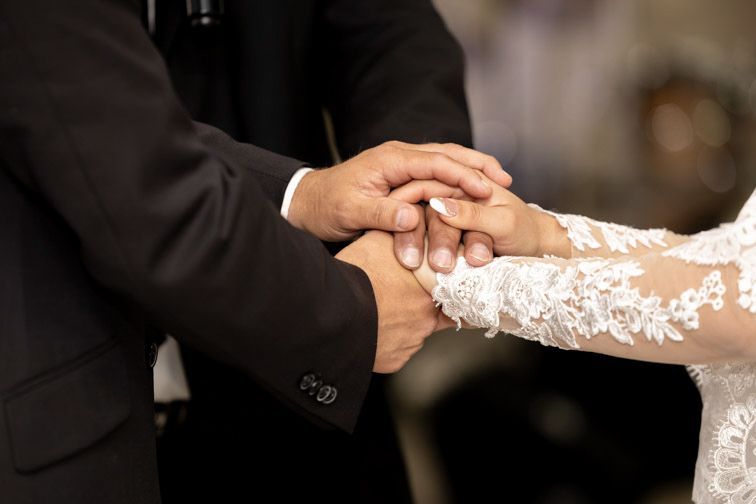Close-up of a groom in a dark suit clasping the hands of a bride in a white lace sleeve during a wedding ceremony.