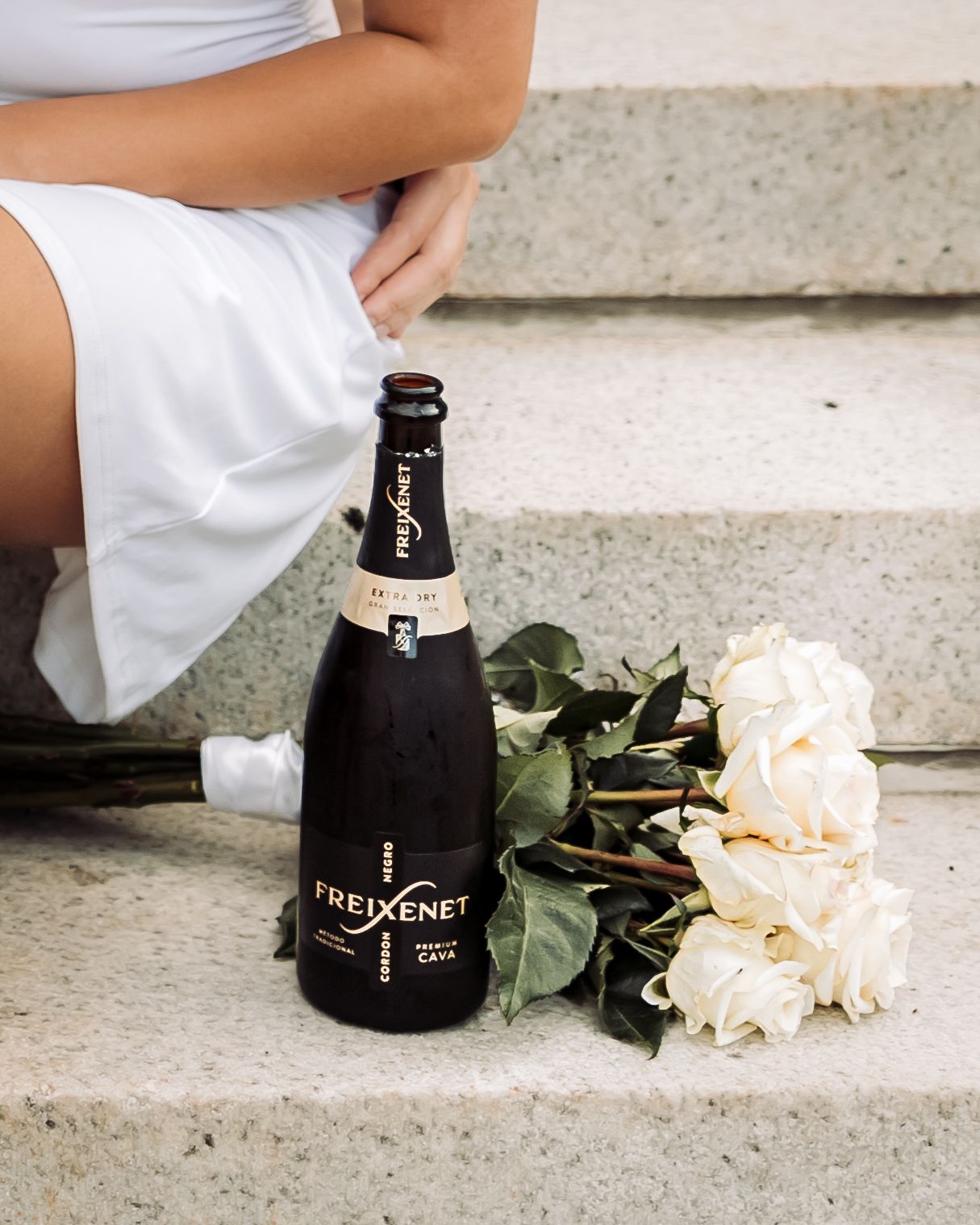Freixenet Cava bottle on stone steps beside white roses with a woman in a white dress.