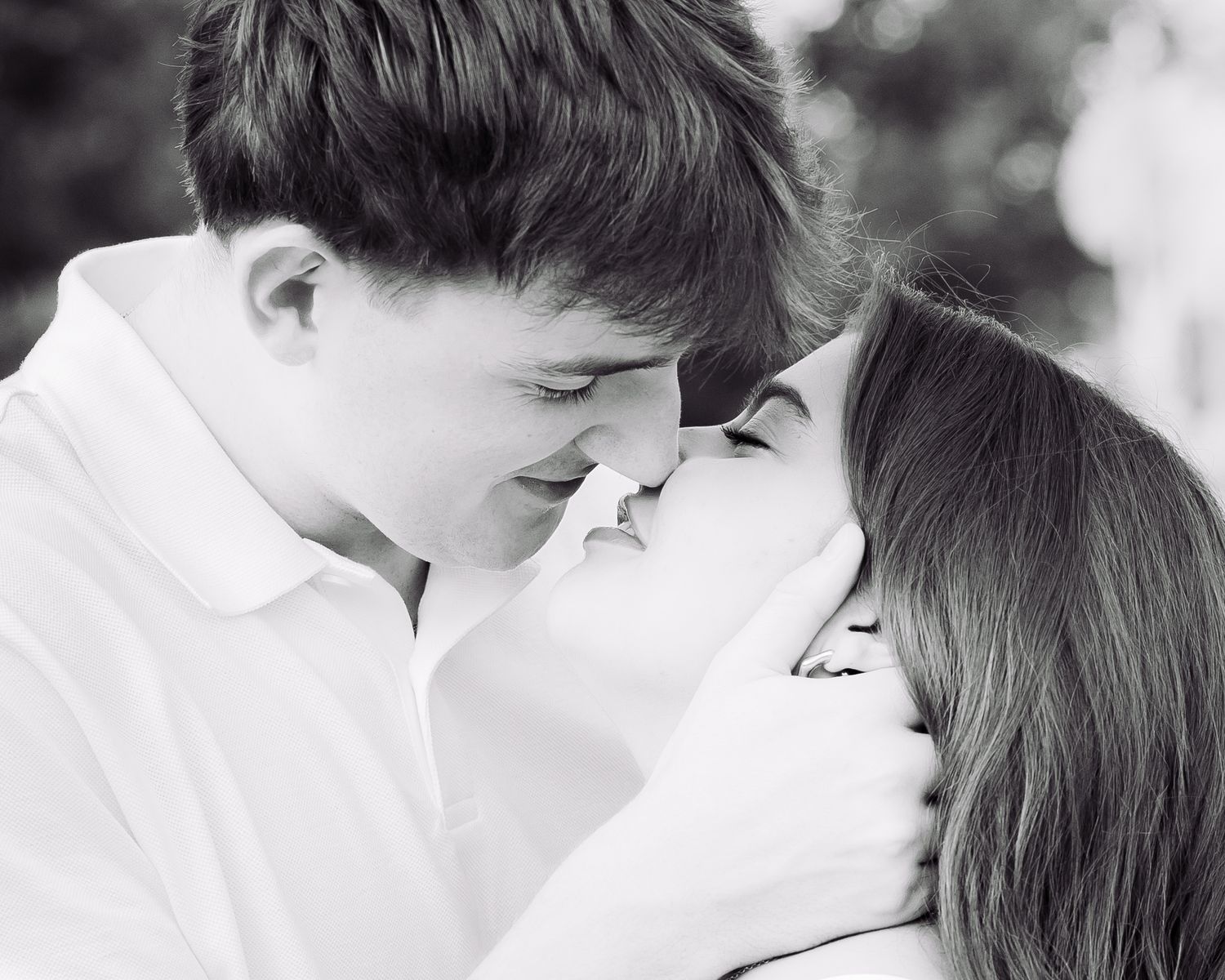 Black and white photo of a young couple sharing a romantic almost-kiss, noses touching tenderly outdoors.