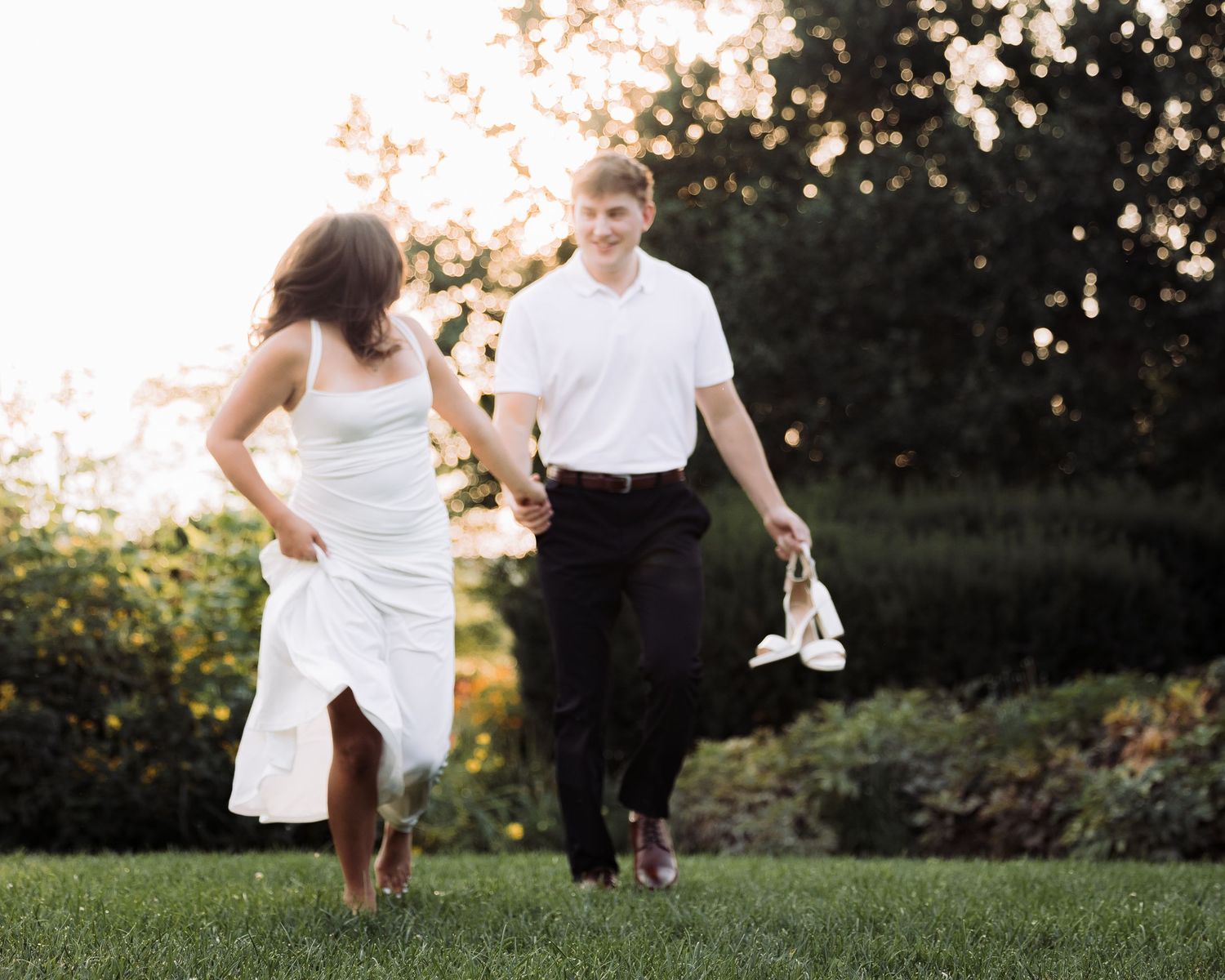 Couple in white dress and black pants holding shoes while running through lush green garden at golden hour.