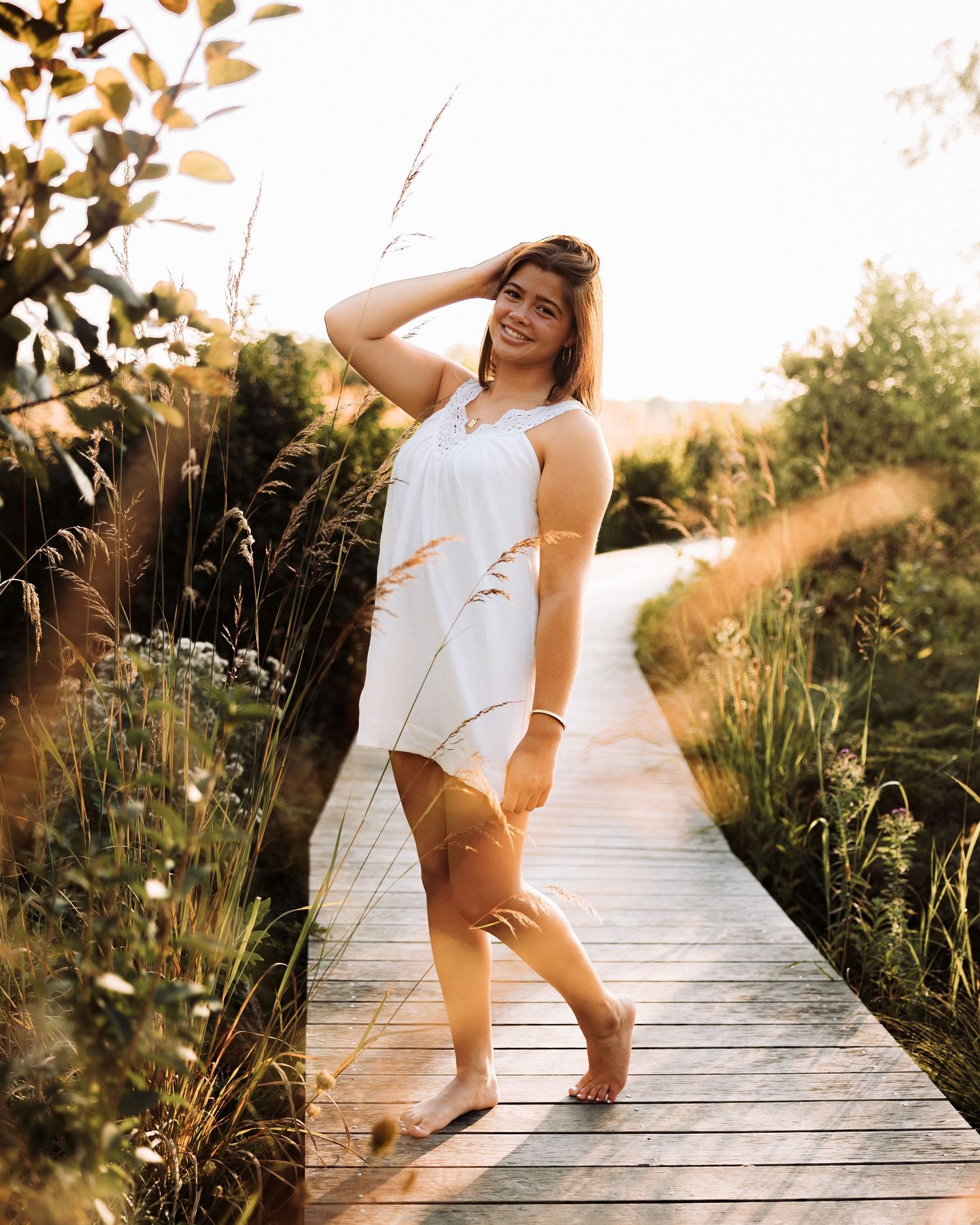 Young woman in white dress smiling on wooden boardwalk path surrounded by tall golden grasses at sunset.