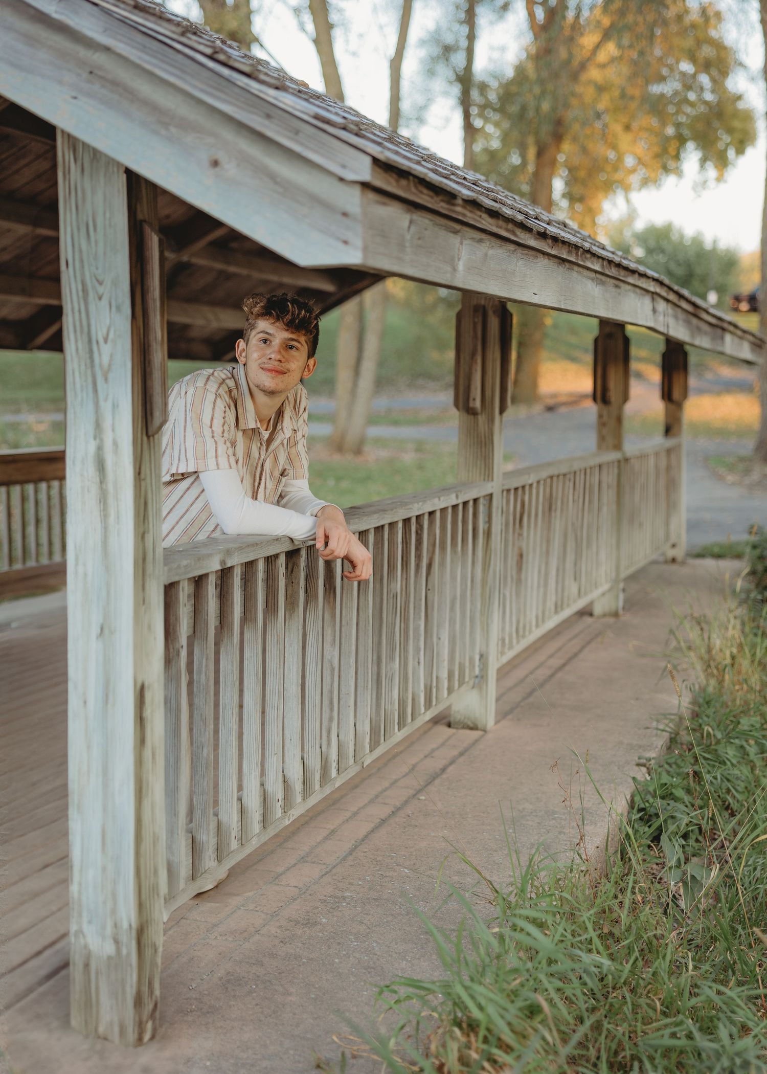 Young person leaning on wooden railed bridge in rural park setting during golden hour.