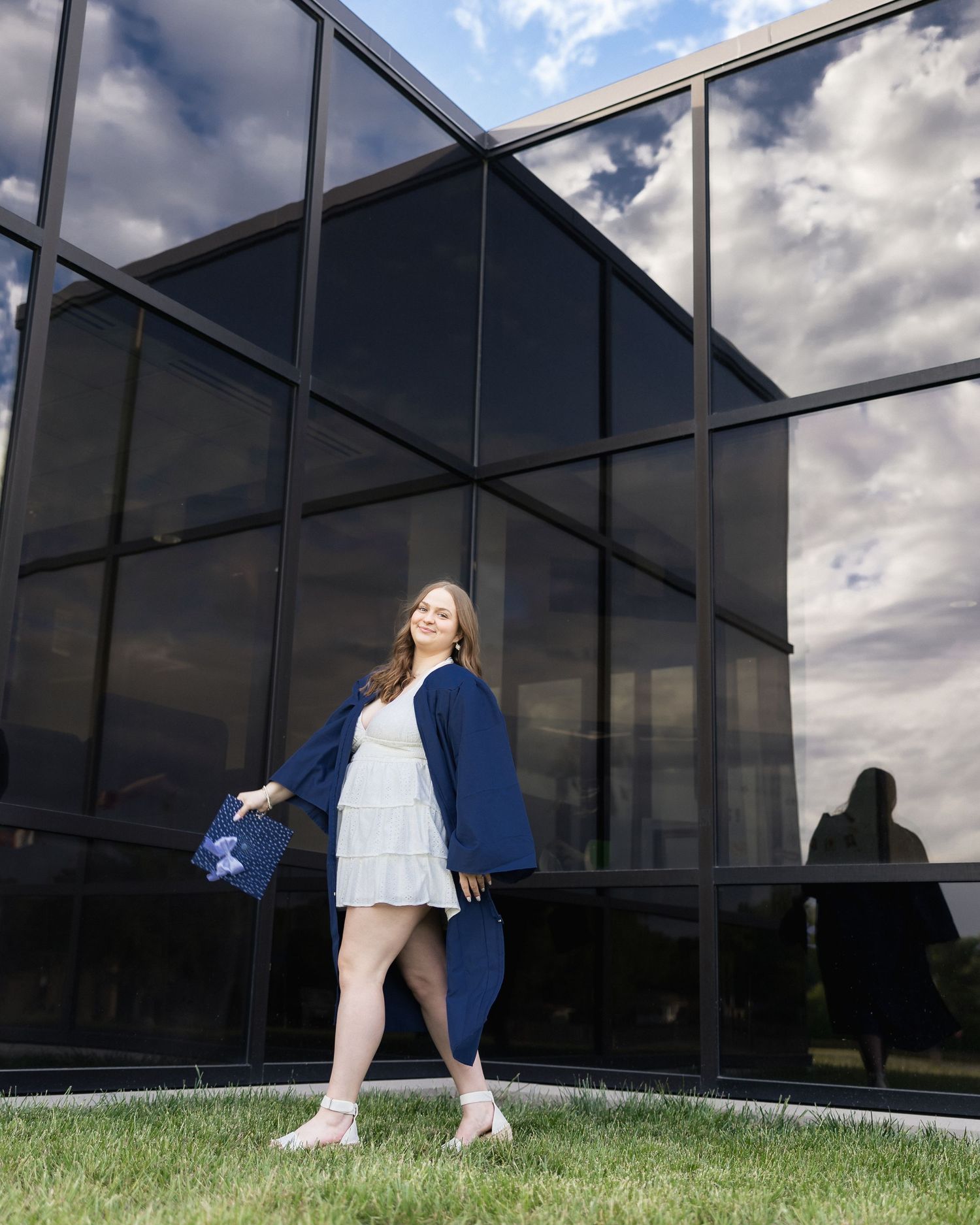 Young woman in white dress and navy graduation gown poses on grass before modern glass building.