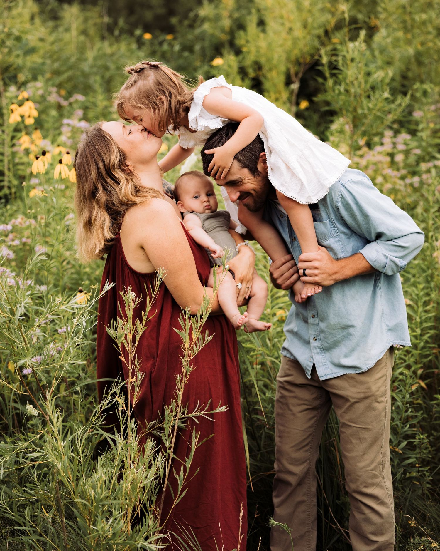 A joyful family of four shares a tender moment in a lush wildflower meadow, parents kissing their toddler while holding a newborn.