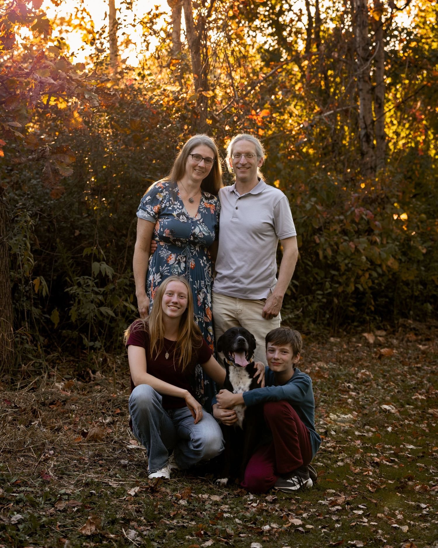 Family of four with a black and white dog posing together outdoors among colorful autumn trees in warm golden sunset light.