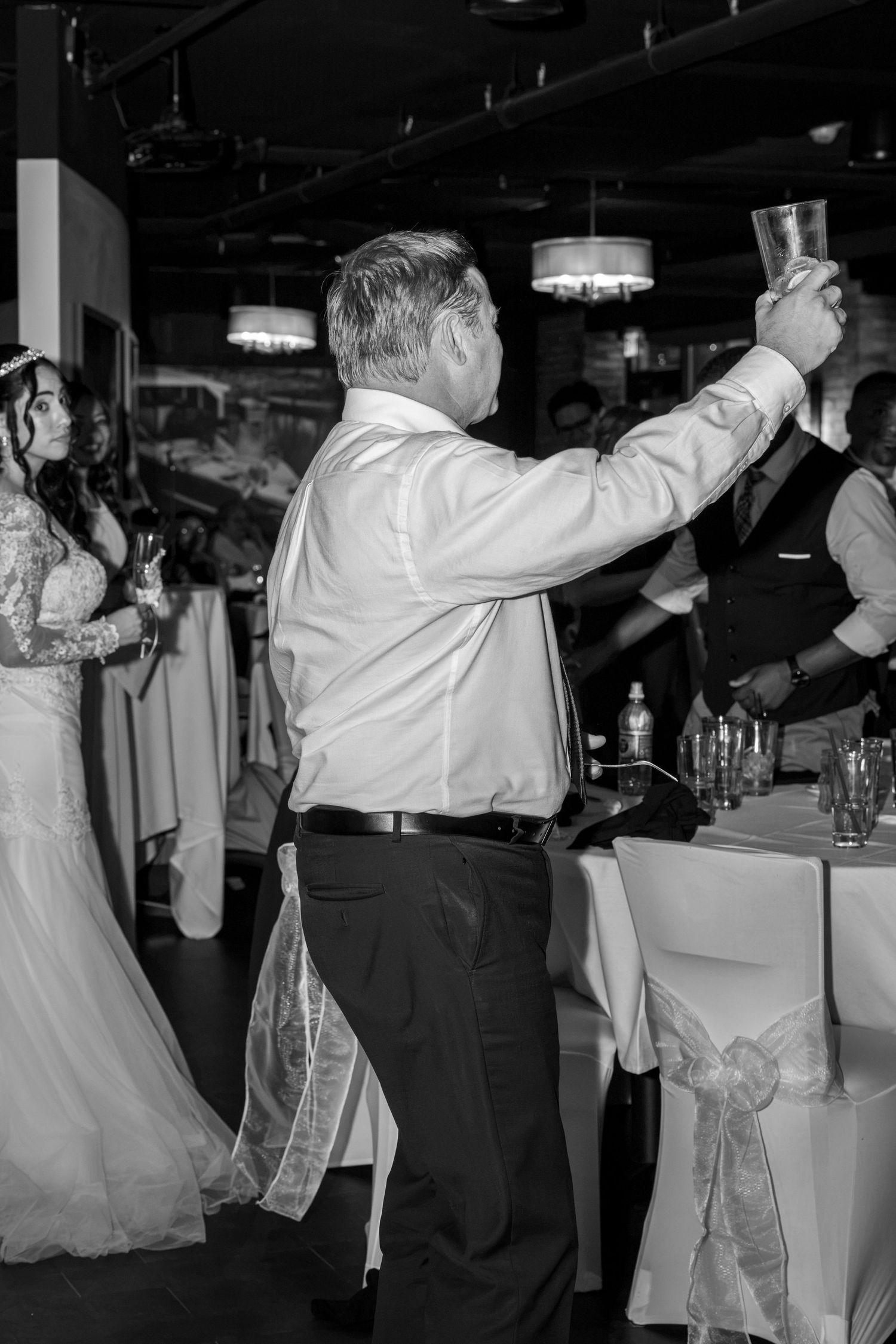 Black and white photo of a man raising a glass in a toast at a wedding reception while a bride in a lace gown stands nearby.
